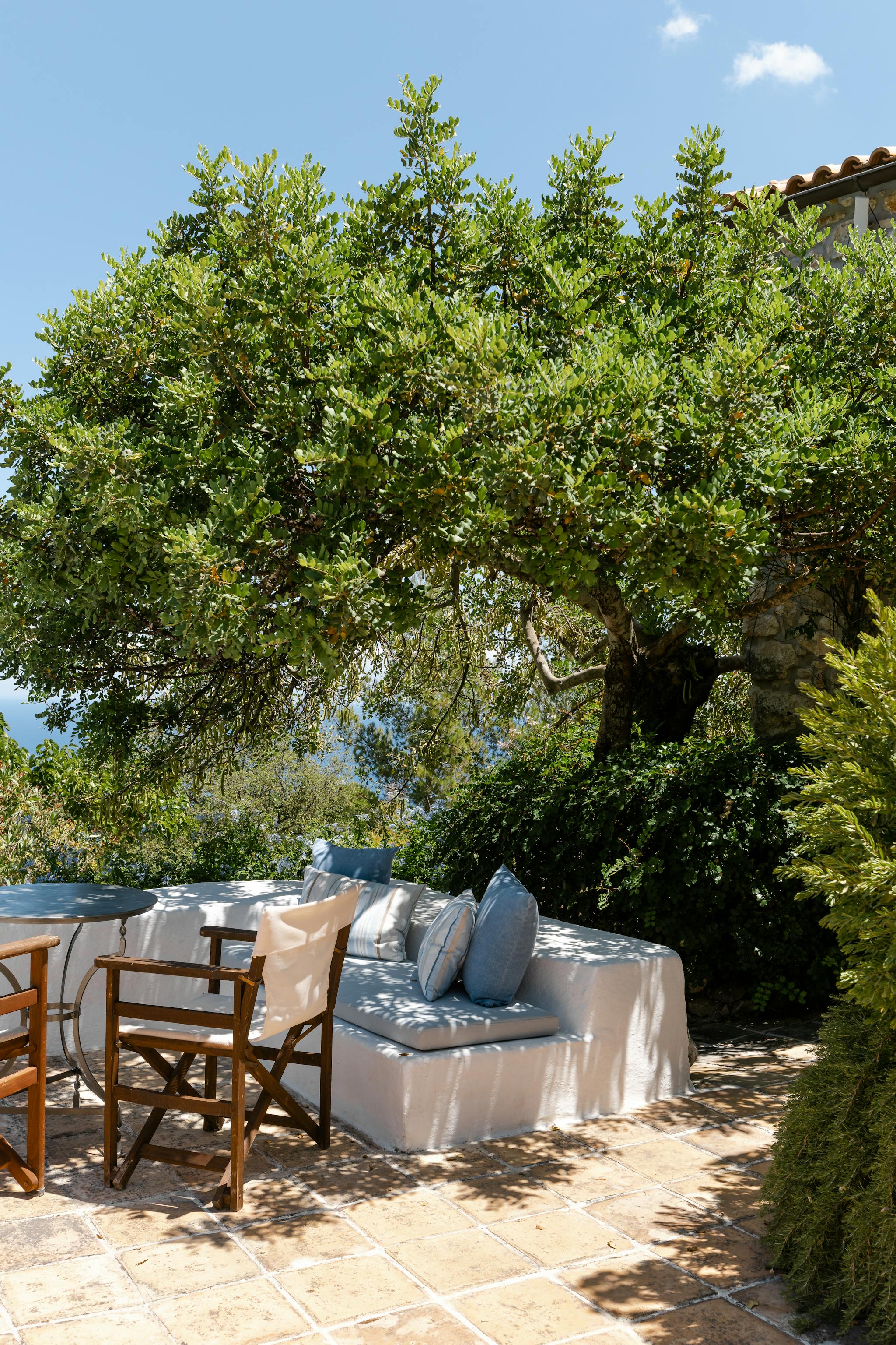 Shaded terrace seating with white cushioned sofa and wooden chairs beneath mature tree canopy. Dappled sunlight creates peaceful garden retreat with glimpse of red-tiled roof.