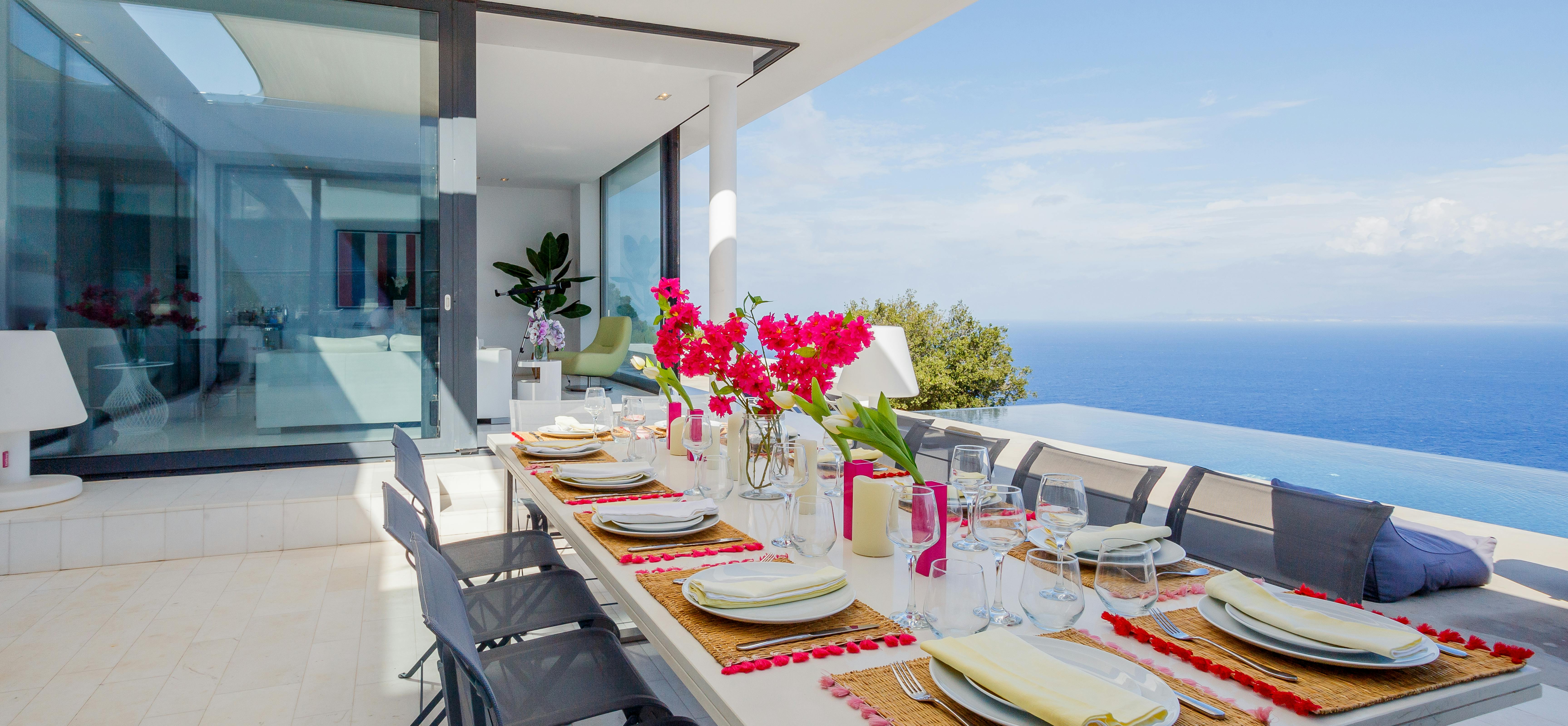 Outdoor dining table set with colourful linens and pink bougainvillea centrepiece overlooking infinity pool and endless sea. Modern white villa with floor-to-ceiling glass doors frames the luxurious alfresco setting.