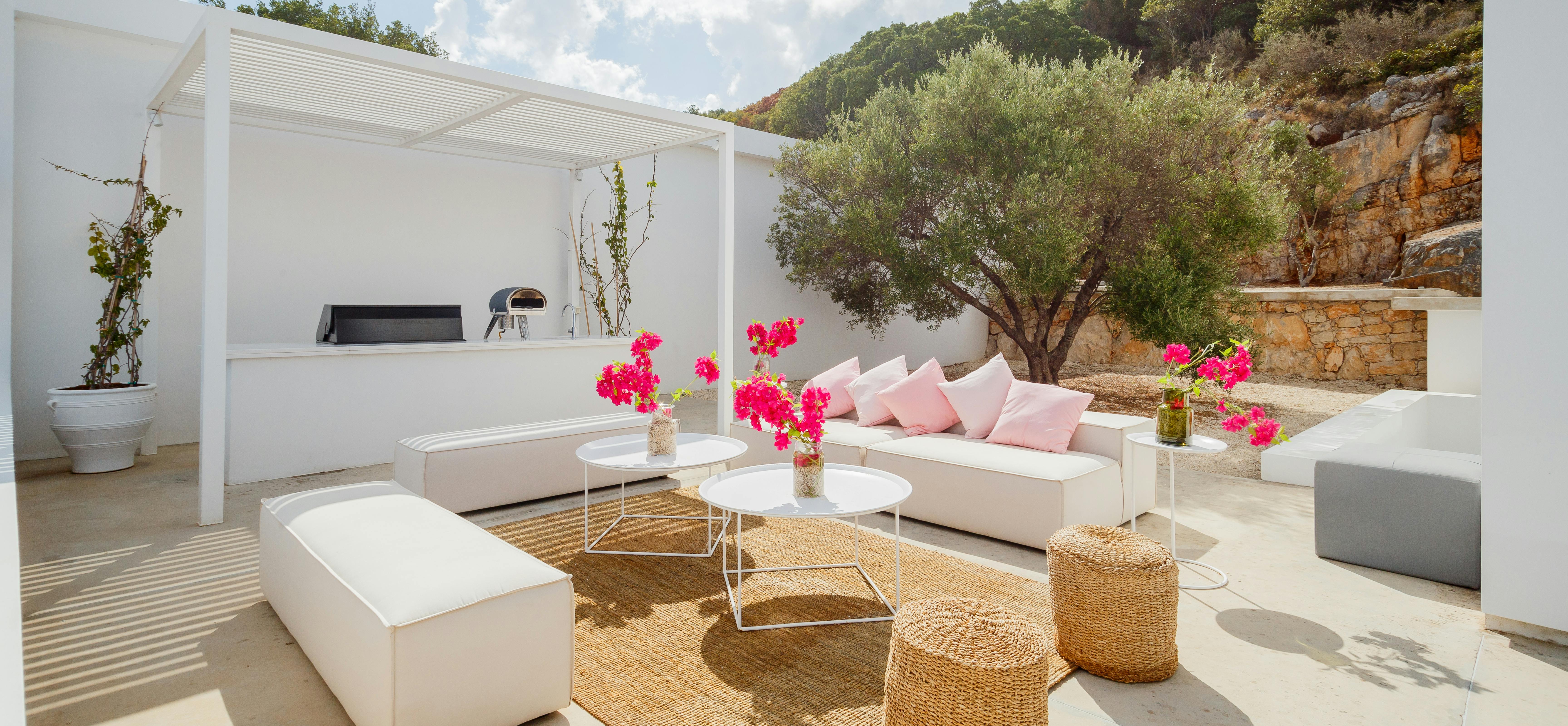 White outdoor living room with built-in kitchen bar, pink-cushioned sofas, and natural fiber accents beneath white pergola. Ancient olive tree and stone hillside provide dramatic natural backdrop.