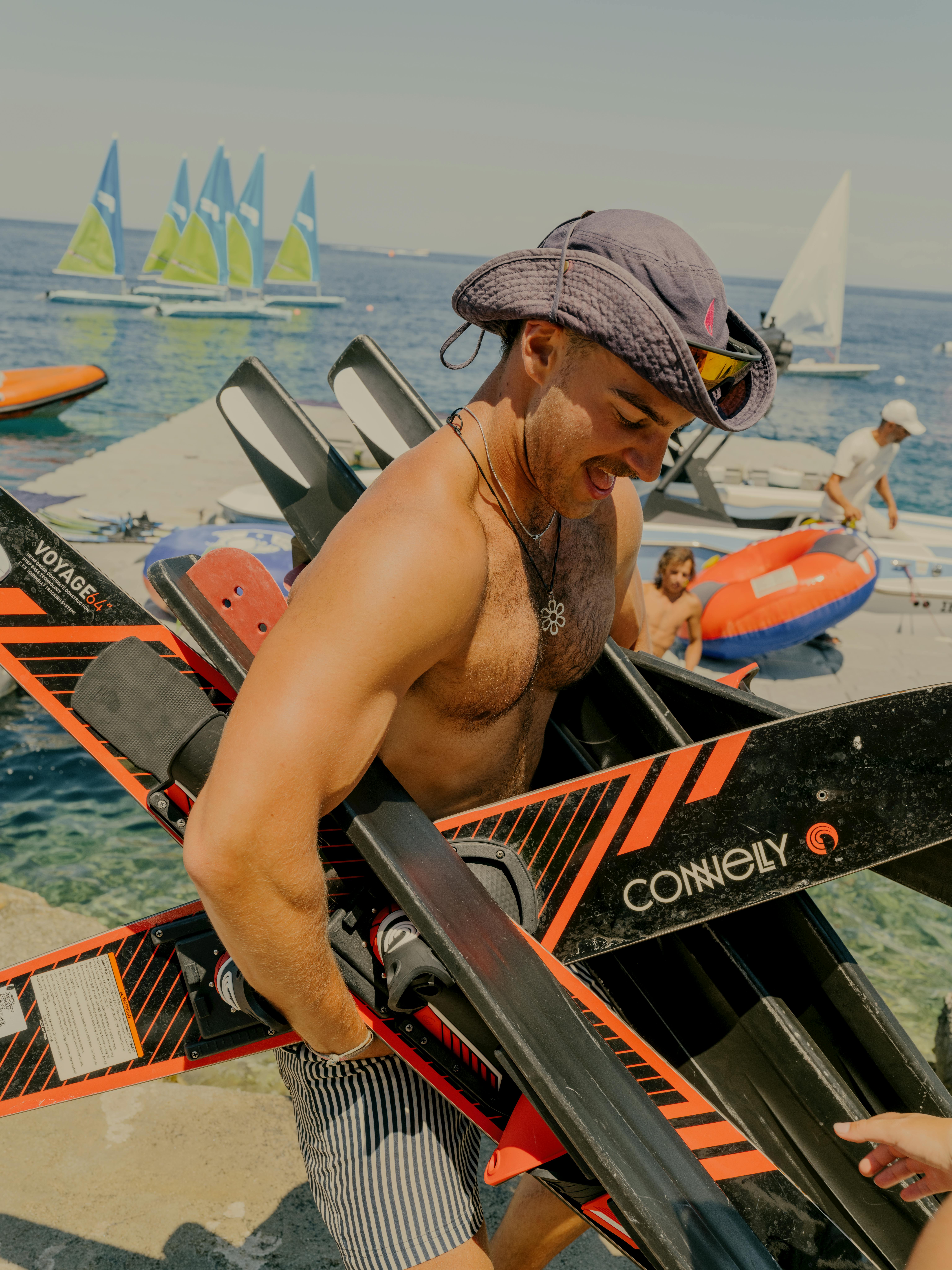 A smiling man wearing a blue safari boonie hat and striped swim shorts carries a black and orange Connelly wakeboard while standing on a dock. He wears sunglasses on his hat and a necklace. Behind him, clear turquoise water is filled with activity - small sailboats with yellow and blue sails, people on inflatable tubes, and various watercraft. A busy beach or water sports area stretches into the background under hazy skies.