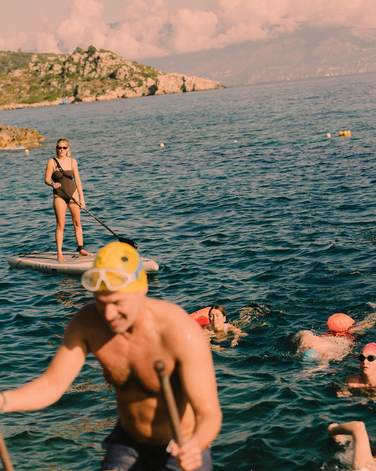 A group enjoying a golden-hour swim in deep blue water. In the foreground, a person wearing a yellow snorkel mask is partially visible and out of focus. Behind them, a woman in a black swimsuit and sunglasses stands on a paddleboard holding a paddle, while several people swim in the water wearing orange flotation devices and snorkel gear. In the background, rocky coastal hills covered with scrub vegetation rise from the water under dramatic pink-tinged clouds at sunset.