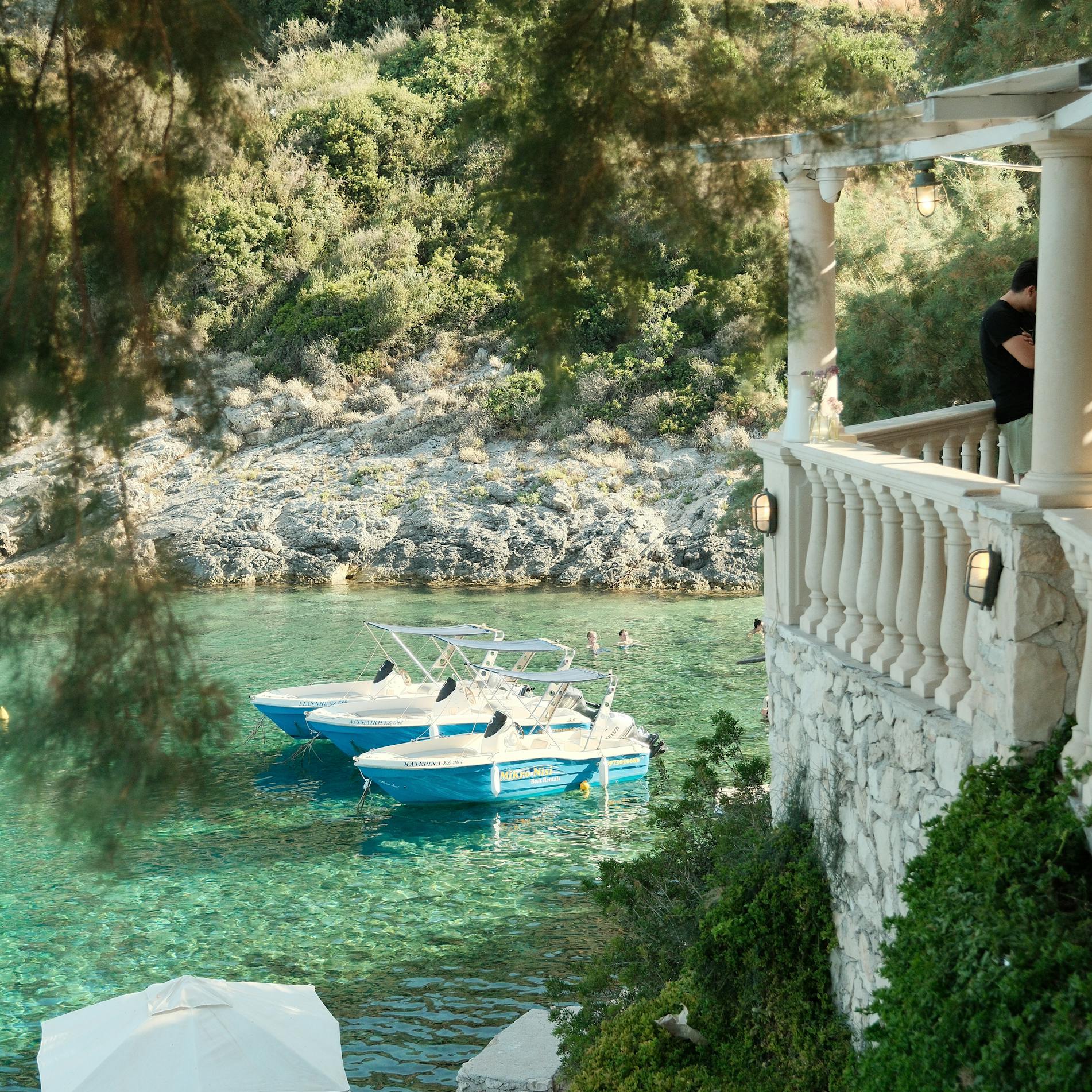 A serene cove with crystal-clear turquoise water, viewed from a white ornate balcony with classical balustrades. Two small boats are moored in the protected inlet, surrounded by a rocky shoreline covered in dense green vegetation and trees. Below, a narrow concrete dock extends into the water where people can be seen relaxing under a white umbrella. The hillside behind is rugged and covered with Mediterranean scrub and pine trees, creating a peaceful, secluded atmosphere.