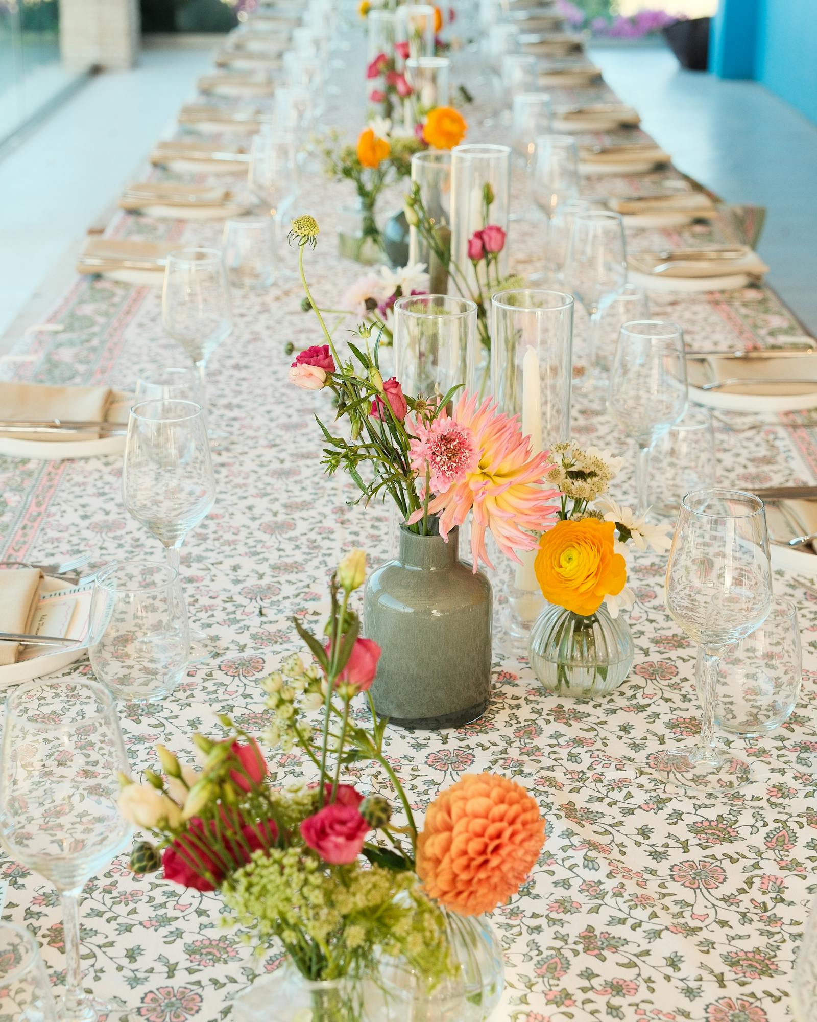 A long dining table set for an elegant outdoor event, photographed lengthwise to show the repeating pattern of place settings stretching into soft focus. The table is covered with a delicate floral-patterned tablecloth in soft pastels. Down the center, a series of small floral arrangements in varied vases feature cheerful blooms - coral dahlias, bright yellow ranunculus, pink spray roses, and wildflowers in muted ceramic vessels and clear glass bottles. Each place setting includes natural woven chargers, folded napkins, and multiple wine glasses. Tall cylinder vases with single stems create vertical interest between the lower arrangements. The bright, airy setting with white walls and colorful flowers visible in the blurred background creates a fresh, summery Mediterranean garden party atmosphere.