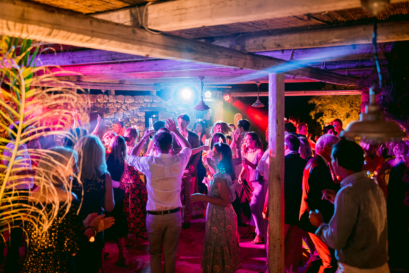 A lively nighttime wedding reception dance floor packed with guests celebrating under a rustic wooden pergola. Dramatic colored lighting bathes the crowd in vibrant pink, purple, blue, and red hues, with stage lights and spotlights creating dynamic beams through the space. Wedding guests with arms raised dance energetically in formal and semi-formal attire. The open-air venue features exposed wooden beams overhead, stone walls in the background, and decorative plants including palm fronds on the left. Trees illuminated by uplighting are visible in the darkness beyond the structure. The DJ or band equipment can be seen against the far wall. The electric atmosphere captures the joy and energy of the late-night celebration.