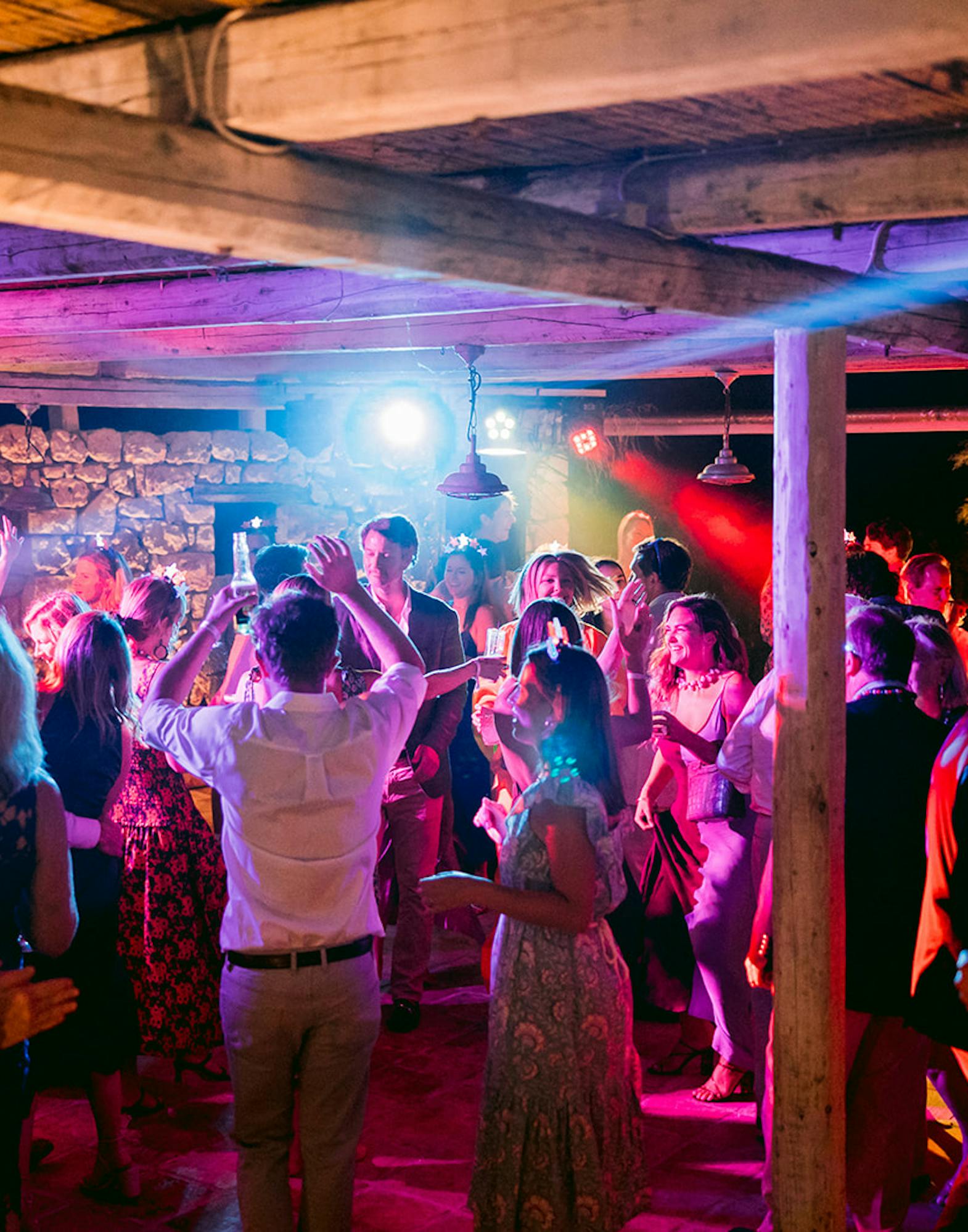 A lively nighttime wedding reception dance floor packed with guests celebrating under a rustic wooden pergola. Dramatic colored lighting bathes the crowd in vibrant pink, purple, blue, and red hues, with stage lights and spotlights creating dynamic beams through the space. Wedding guests with arms raised dance energetically in formal and semi-formal attire. The open-air venue features exposed wooden beams overhead, stone walls in the background, and decorative plants including palm fronds on the left. Trees illuminated by uplighting are visible in the darkness beyond the structure. The DJ or band equipment can be seen against the far wall. The electric atmosphere captures the joy and energy of the late-night celebration.