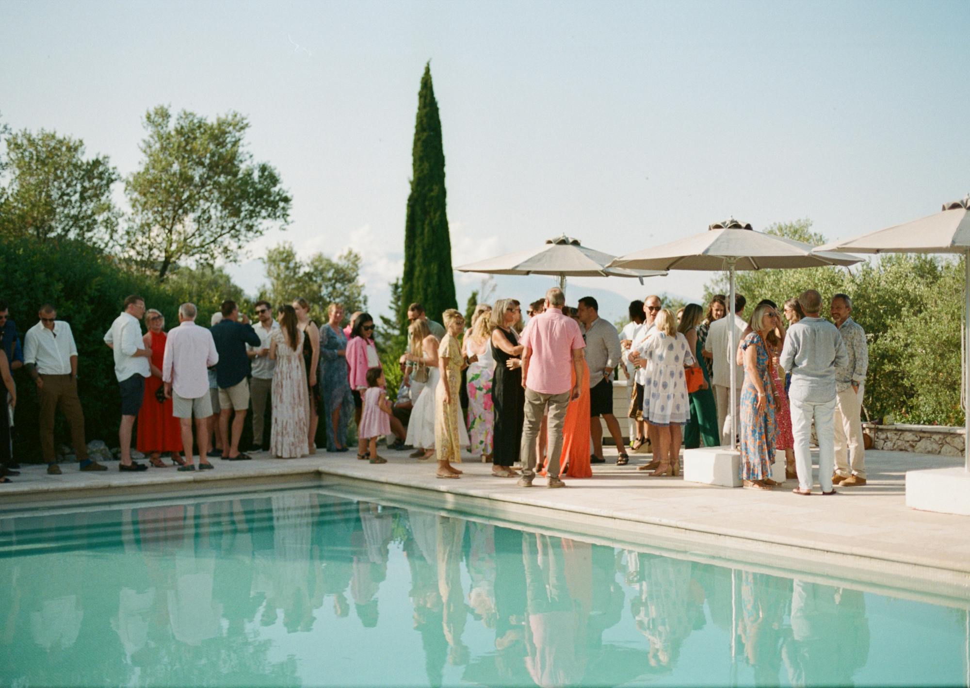 An elegant outdoor cocktail reception beside a pristine turquoise swimming pool on a warm afternoon. Guests in smart casual and semi-formal summer attire mingle on the pale stone pool deck, their figures and the tall beige market umbrellas reflected in the still water. The crowd includes adults and children wearing colorful sundresses, linen shirts, and light trousers in shades of pink, coral, white, and floral patterns. A distinctive tall Italian cypress tree stands as a vertical accent in the center background, flanked by olive trees and lush Mediterranean vegetation. The refined, relaxed atmosphere and natural setting create a quintessentially Mediterranean garden party scene under soft afternoon light.
