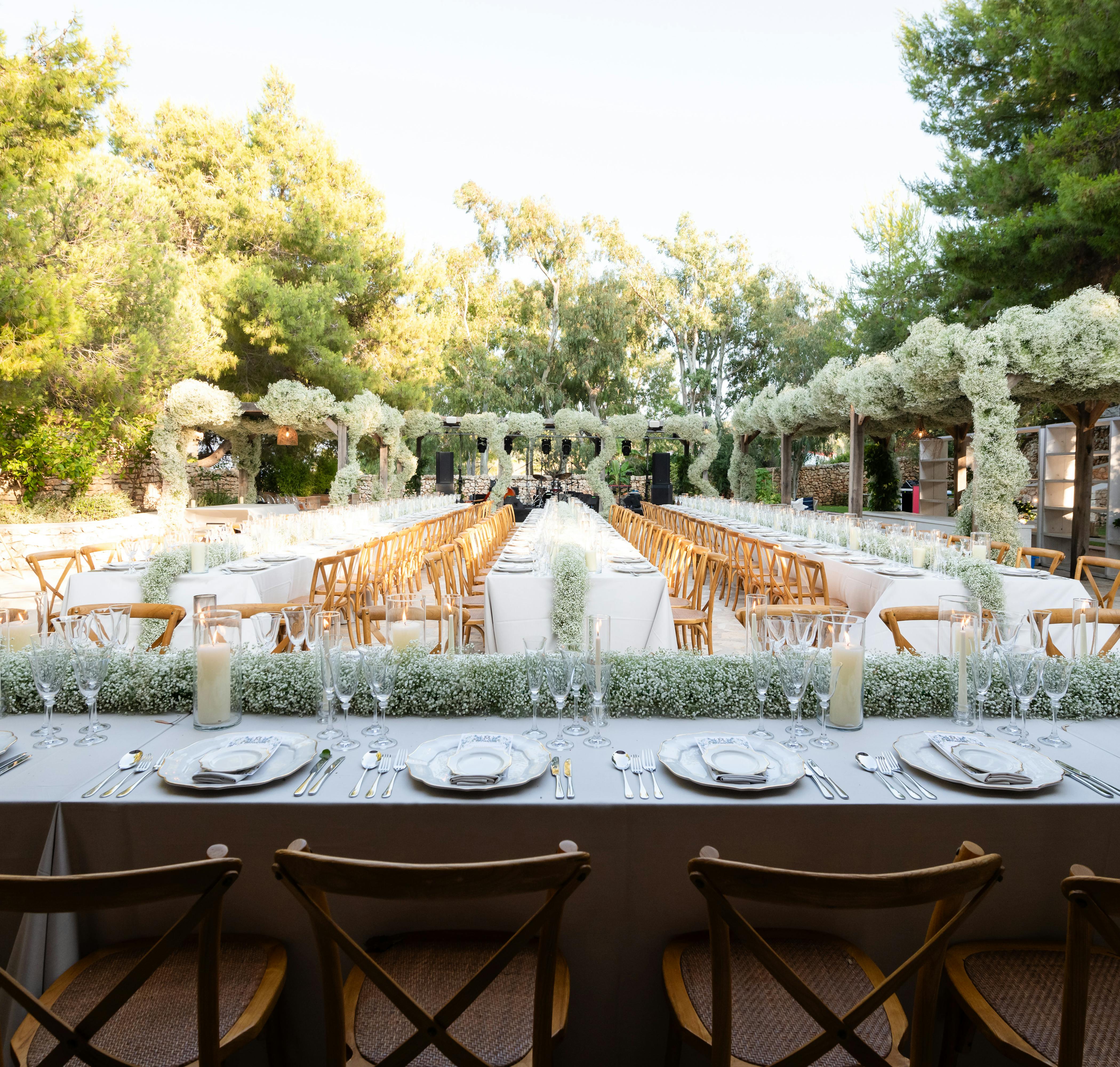An elegant outdoor wedding reception setup photographed from the head table, looking down long rows of pristine white-clothed tables with natural wood cross-back chairs. The sophisticated tablescape features meticulously set place settings with multiple glasses, folded napkins, and gleaming cutlery. A dramatic floral installation creates an overhead canopy - clouds of white baby's breath suspended on an architectural structure forming a tunnel effect down the center aisle. Tall pillar candles in glass cylinders provide additional decoration. Lush greenery surrounds the venue, with mature trees framing the space and a rustic stone building visible in the background. The natural outdoor setting is bathed in soft daylight, creating an refined yet organic Mediterranean garden atmosphere.