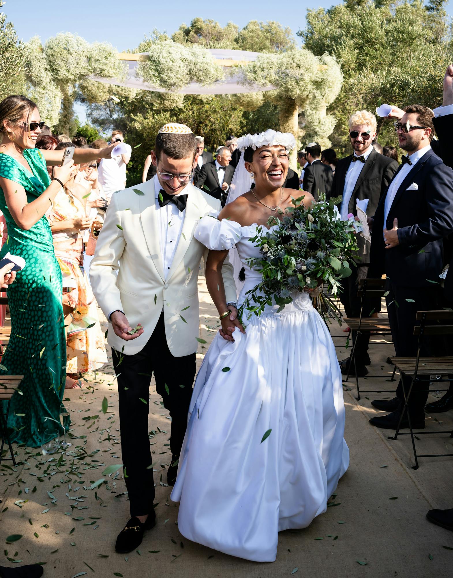 A jubilant recessional moment as the newlyweds walk down the aisle hand-in-hand through an outdoor ceremony space framed by lush olive trees. The groom wears an elegant ivory dinner jacket with black bow tie and trousers, while the beaming bride in a white off-shoulder ball gown with full skirt carries a cascading green foliage bouquet. Wedding guests line both sides of the aisle, waving white programs or napkins in celebration, creating a joyful tunnel of well-wishers. The guests are dressed in formal attire including black tuxedos and elegant gowns in emerald green and other colors. Behind them, a dramatic floral installation covered in white flowers and greenery creates an arbor over the ceremony seating area, with olive groves visible in the background under bright Mediterranean sunlight.