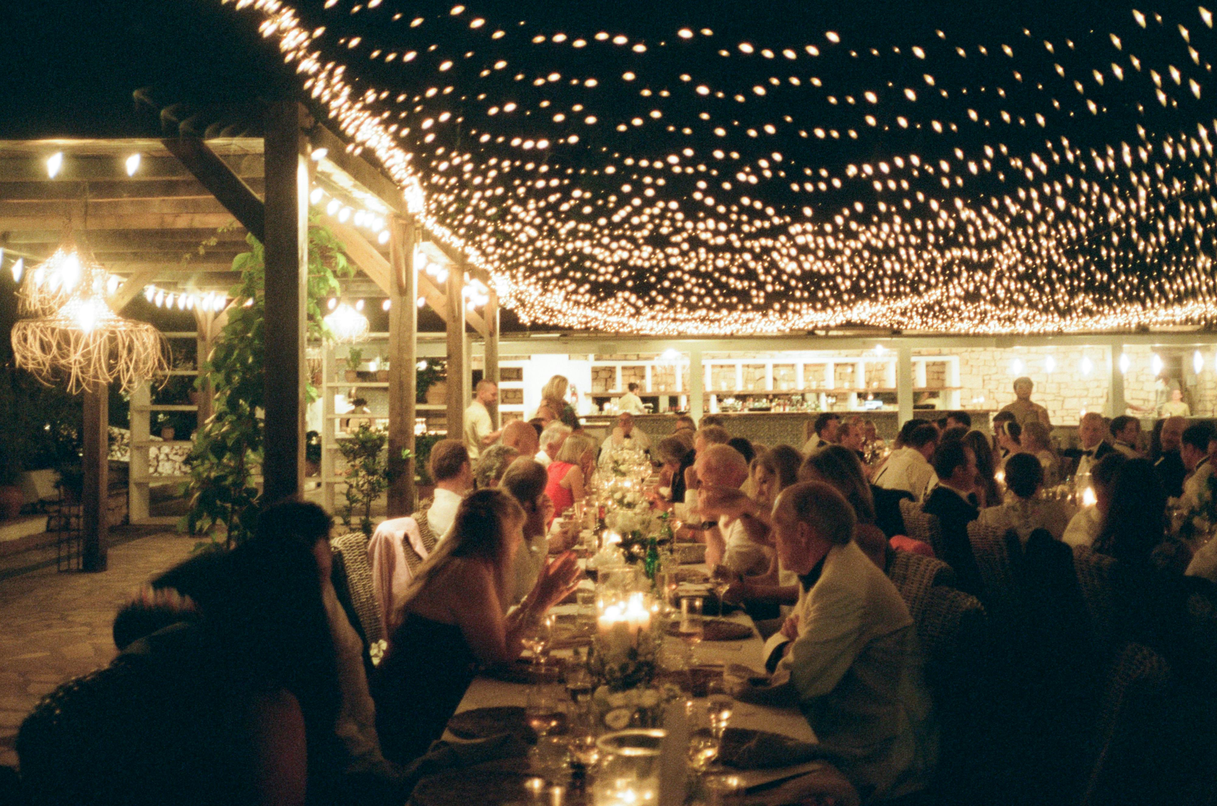 Wedding reception dinner under an elaborate canopy of twinkling string lights. Guests sit at long tables enjoying an evening meal in an open-air venue with white architectural columns and ambient lighting. The overhead fairy lights create a starlit effect across the entire dining space, while floral centerpieces adorn the tables.