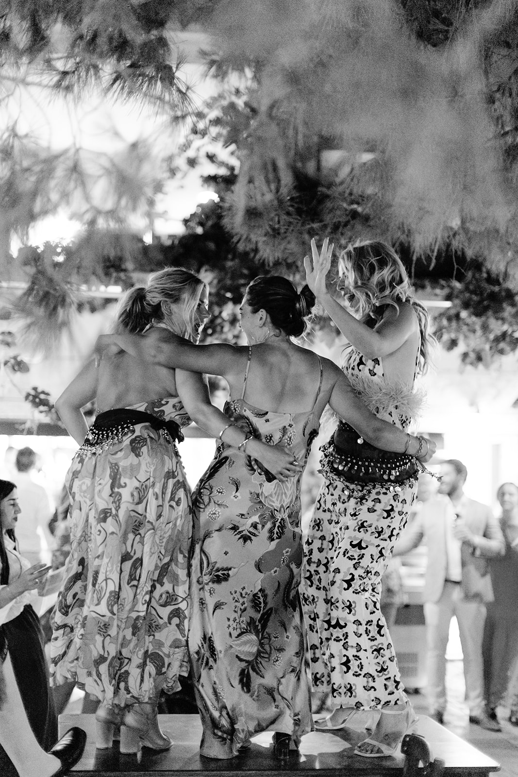 Black and white photo of two women dancing together at an outdoor evening celebration with dramatic smoke or fog effects. They wear patterned dresses and raise their arms joyfully while dancing, with string lights and other guests visible in the atmospheric background.