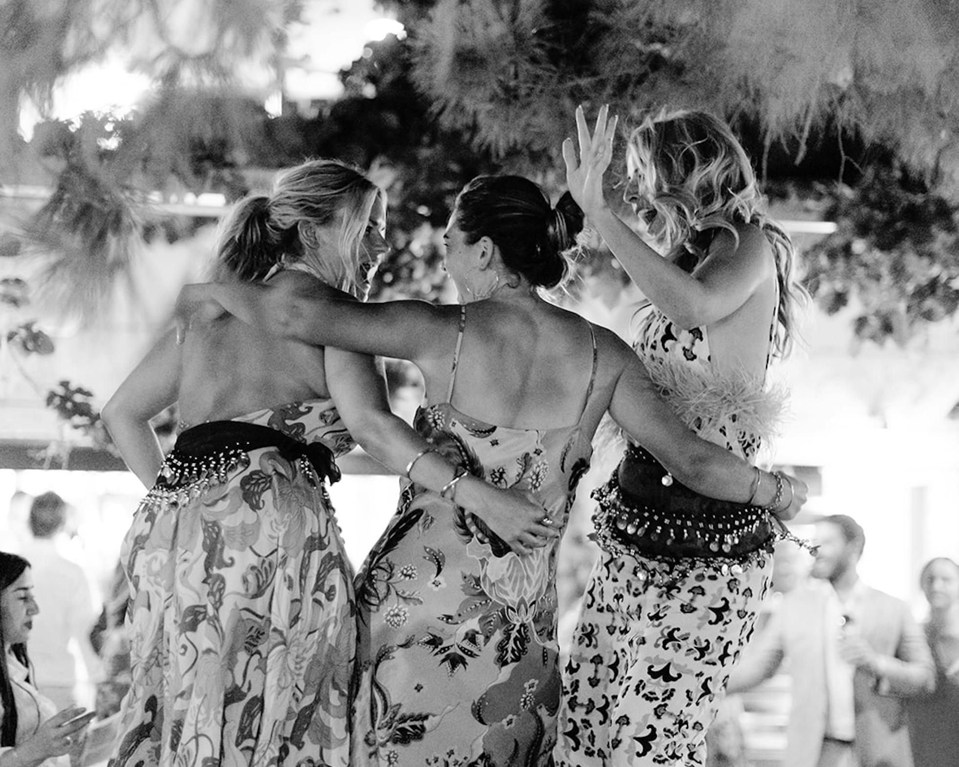 Black and white photo of two women dancing together at an outdoor evening celebration with dramatic smoke or fog effects. They wear patterned dresses and raise their arms joyfully while dancing, with string lights and other guests visible in the atmospheric background.