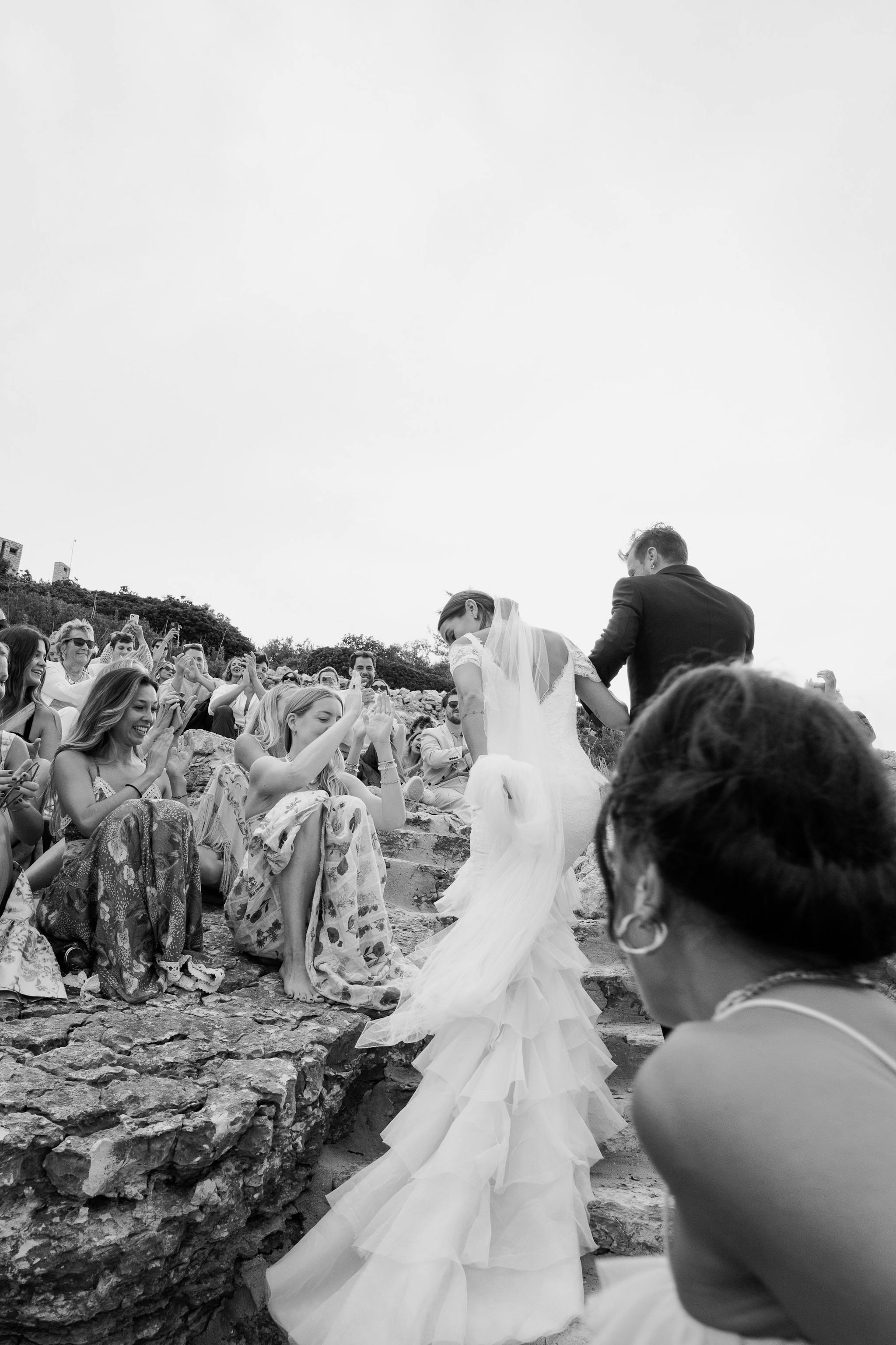 Black and white photo of bride and groom walking down the aisle after their wedding ceremony. The bride's long veil and train flow behind her as guests remain seated on both sides. The ceremony takes place outdoors on natural ground with trees visible in the background.