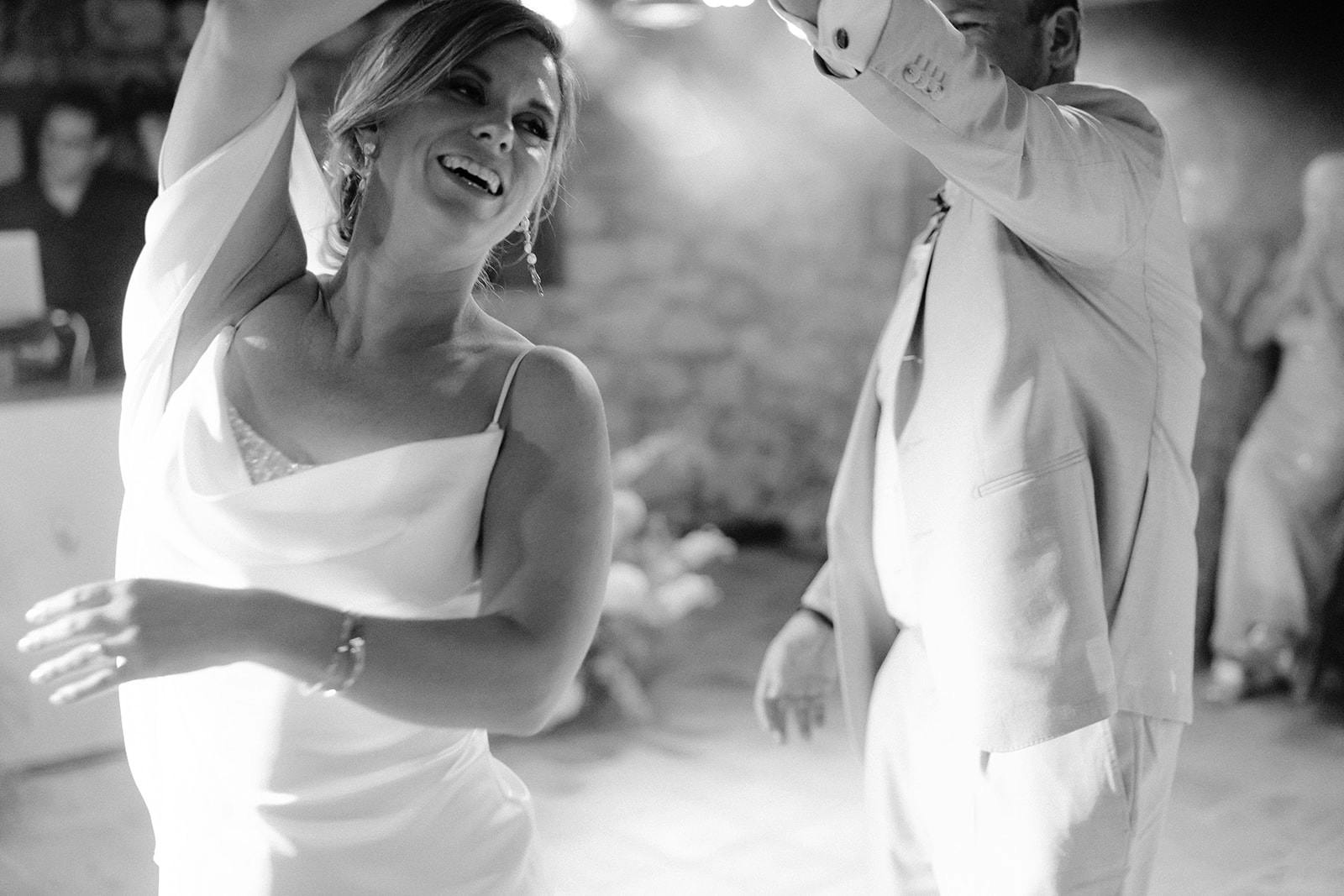 Black and white photo of two brides in white wedding dresses dancing together joyfully at their reception, with one bride laughing radiantly while they hold hands and spin.