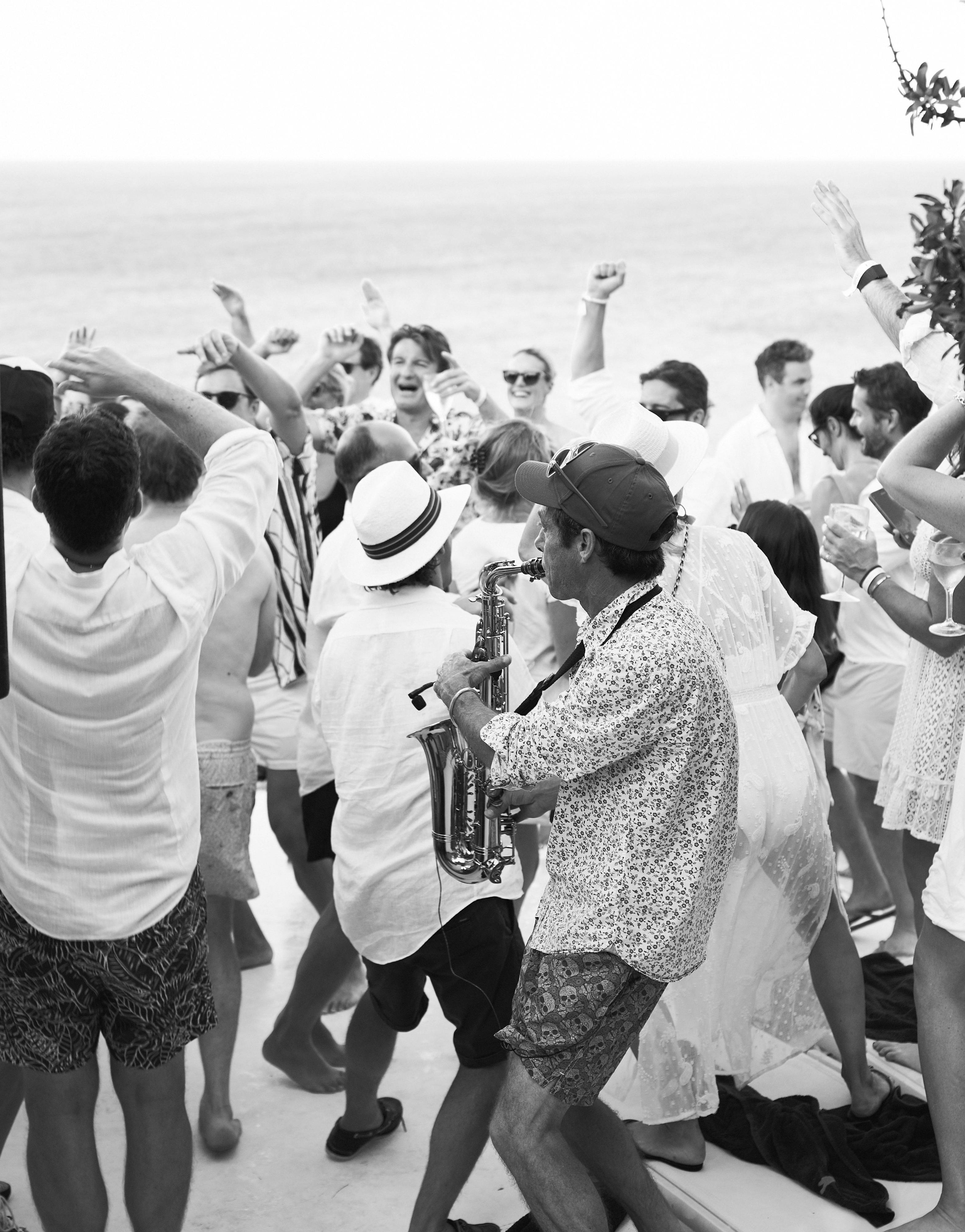 Black and white photo of wedding guests dancing outdoors with live saxophone musician. Guests in summer attire with raised arms dance energetically around a saxophonist in patterned shirt and straw hat, with the ocean visible in the bright, hazy background.