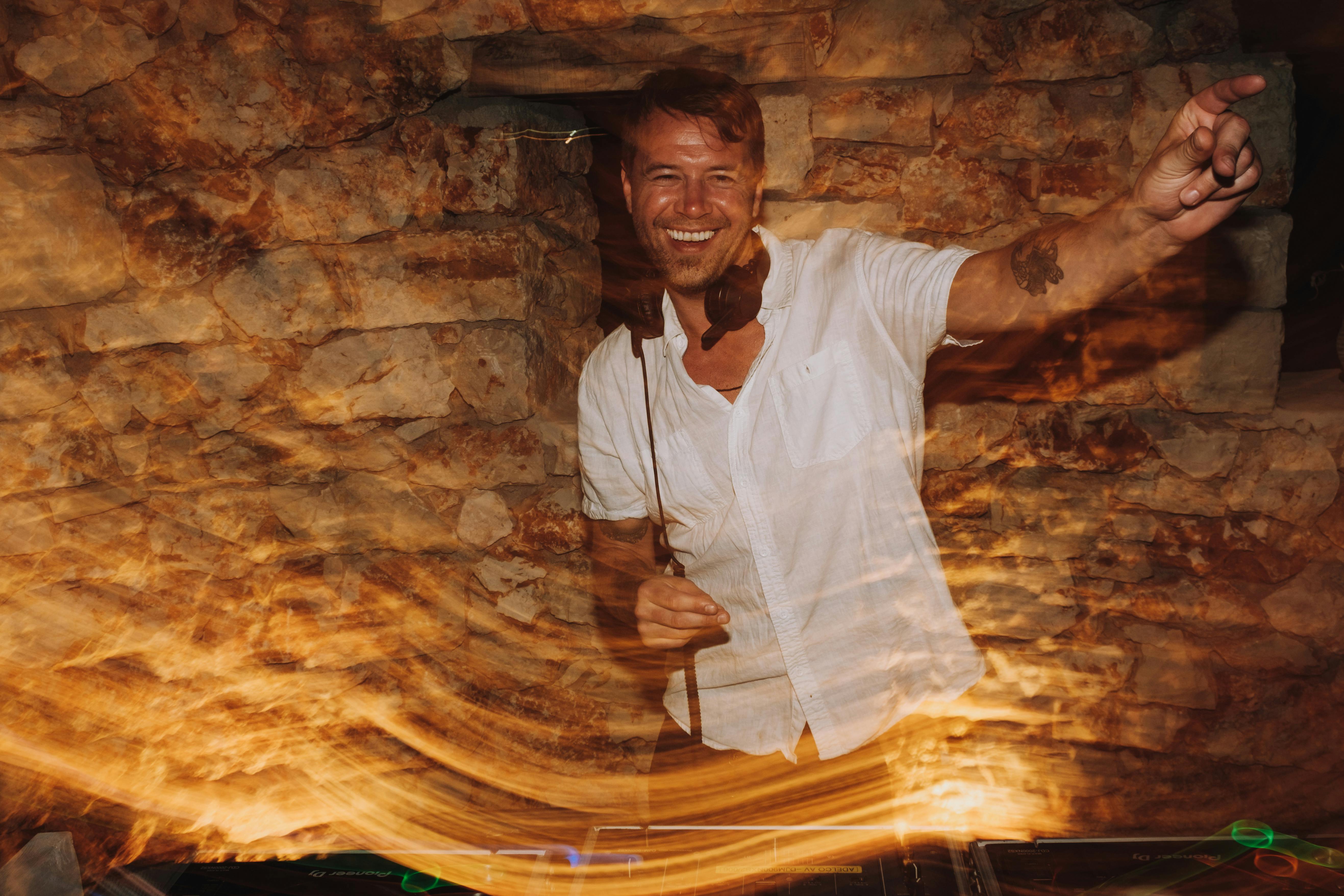 DJ in white shirt performing enthusiastically behind decks with arms raised at night wedding reception. The motion blur effect creates streaks of light across the image, captured against a rustic stone wall backdrop.