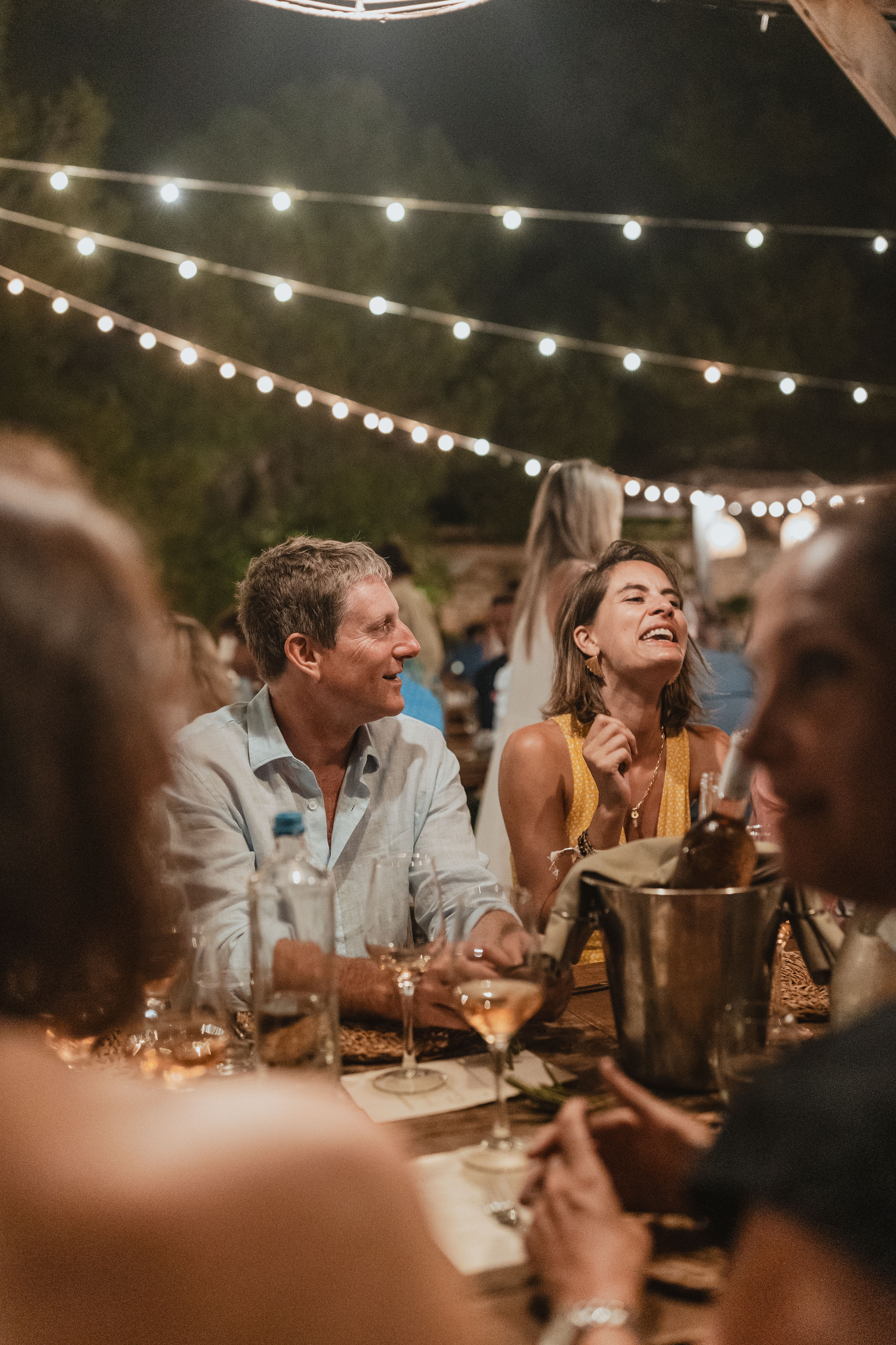 Guests enjoying dinner at an outdoor evening reception under string lights. Guests in casual attire sit at a long table with candles and wine bottles, engaged in conversation and laughter. The warm, intimate atmosphere is created by twinkling overhead lights against the dark night sky.