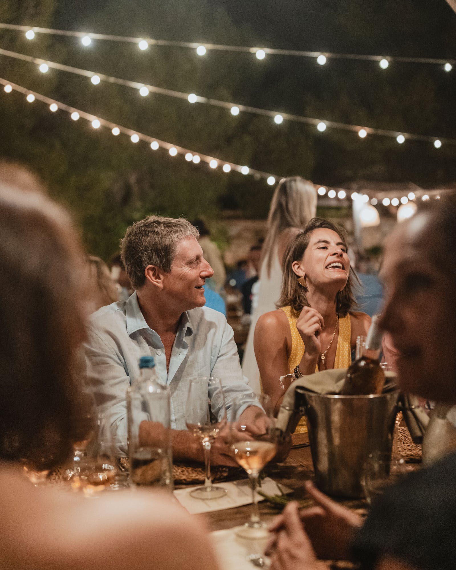 Guests enjoying dinner at an outdoor evening reception under string lights. Guests in casual attire sit at a long table with candles and wine bottles, engaged in conversation and laughter. The warm, intimate atmosphere is created by twinkling overhead lights against the dark night sky.