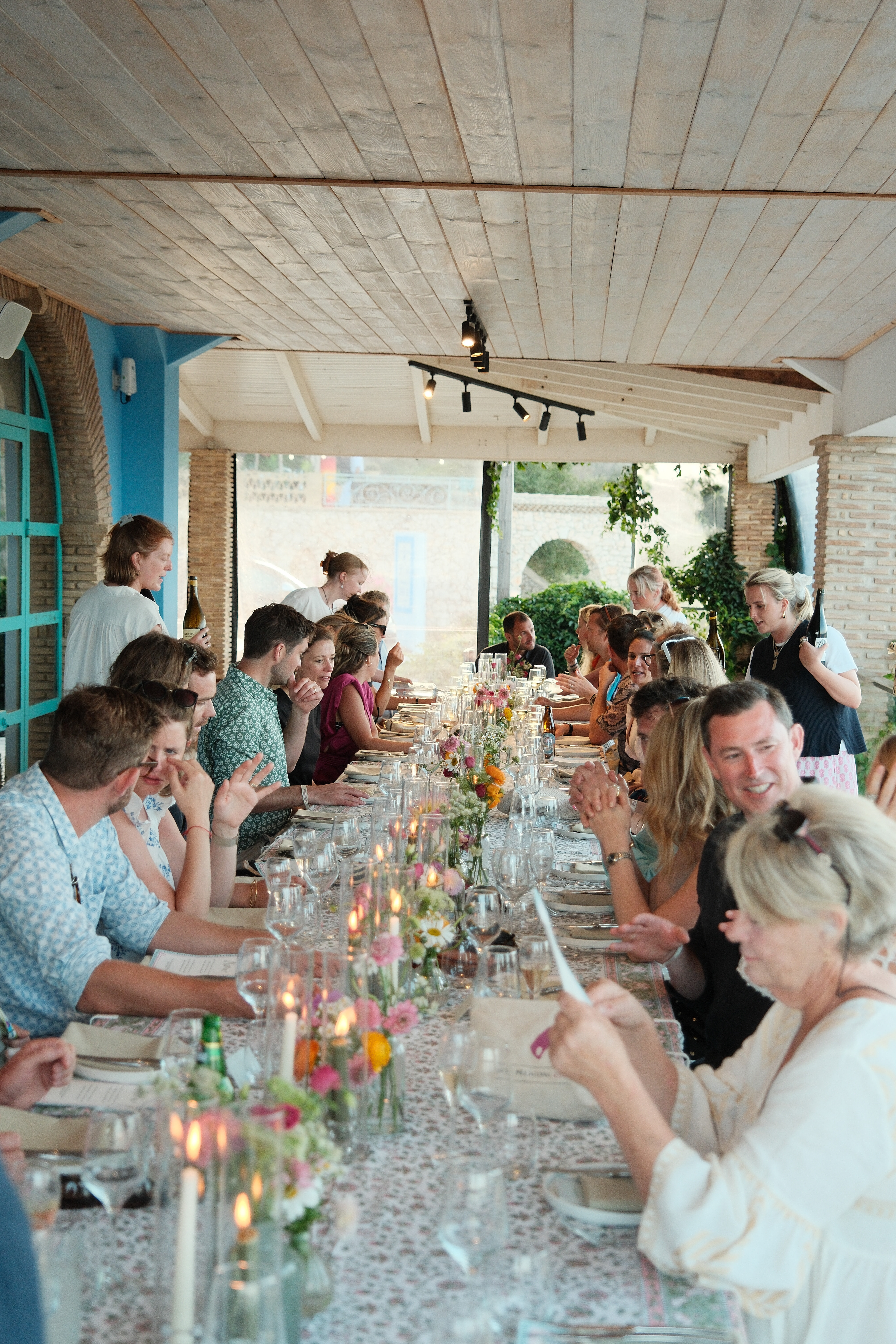 A long communal dining table in a bright, airy restaurant with white-washed wooden ceiling beams and exposed brick columns. Guests in summer attire are seated along both sides of an elegantly decorated table featuring colourful wildflowers, glassware, and candlelight. Natural light streams through, while turquoise accent walls add Mediterranean charm to the intimate dining gathering.