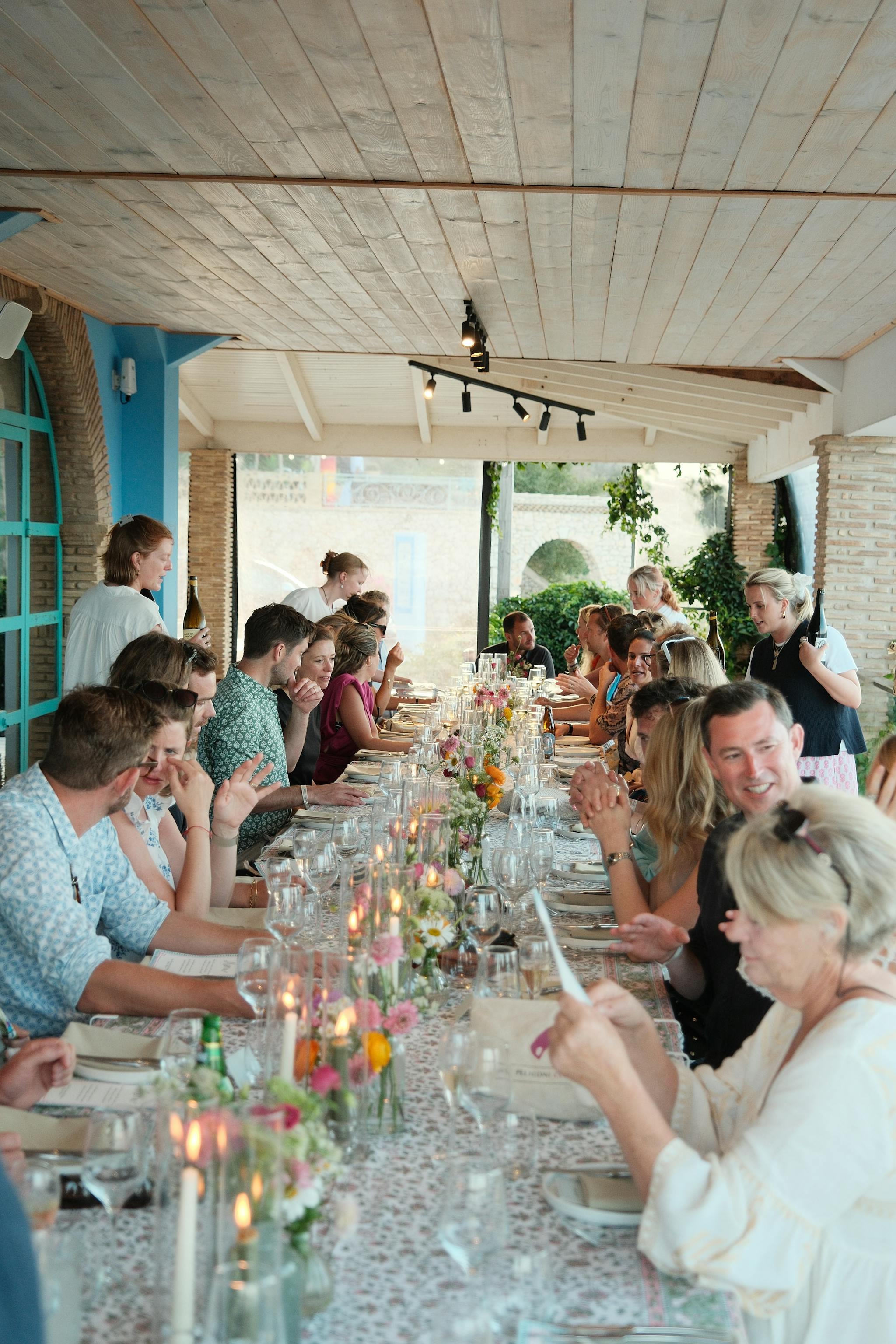 A long communal dining table in a bright, airy restaurant with white-washed wooden ceiling beams and exposed brick columns. Guests in summer attire are seated along both sides of an elegantly decorated table featuring colourful wildflowers, glassware, and candlelight. Natural light streams through, while turquoise accent walls add Mediterranean charm to the intimate dining gathering.