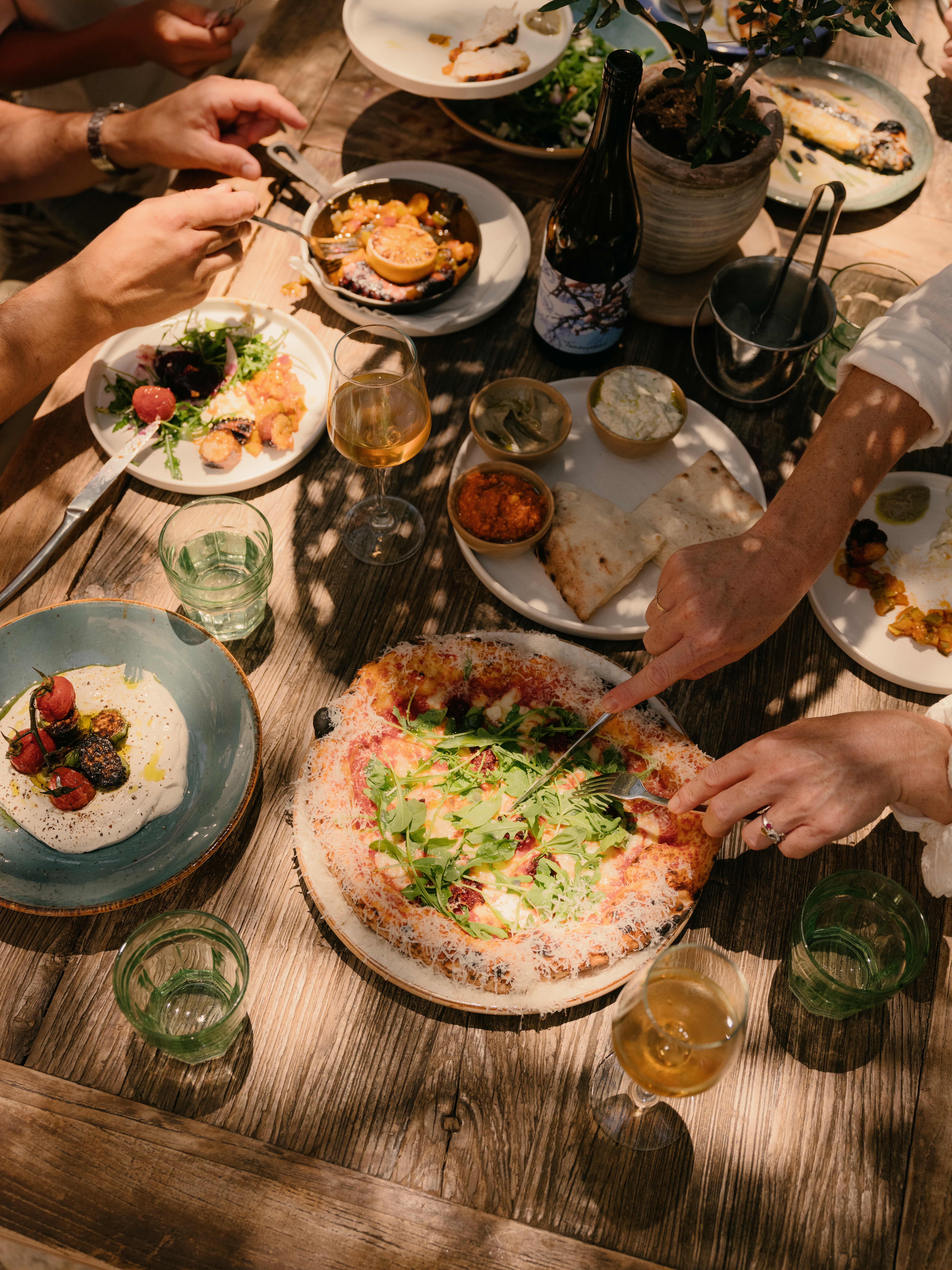 Communal dining table laden with artisan pizza, mezze plates, and wine glasses as hands reach to share food in a warm sun-drenched atmosphere.