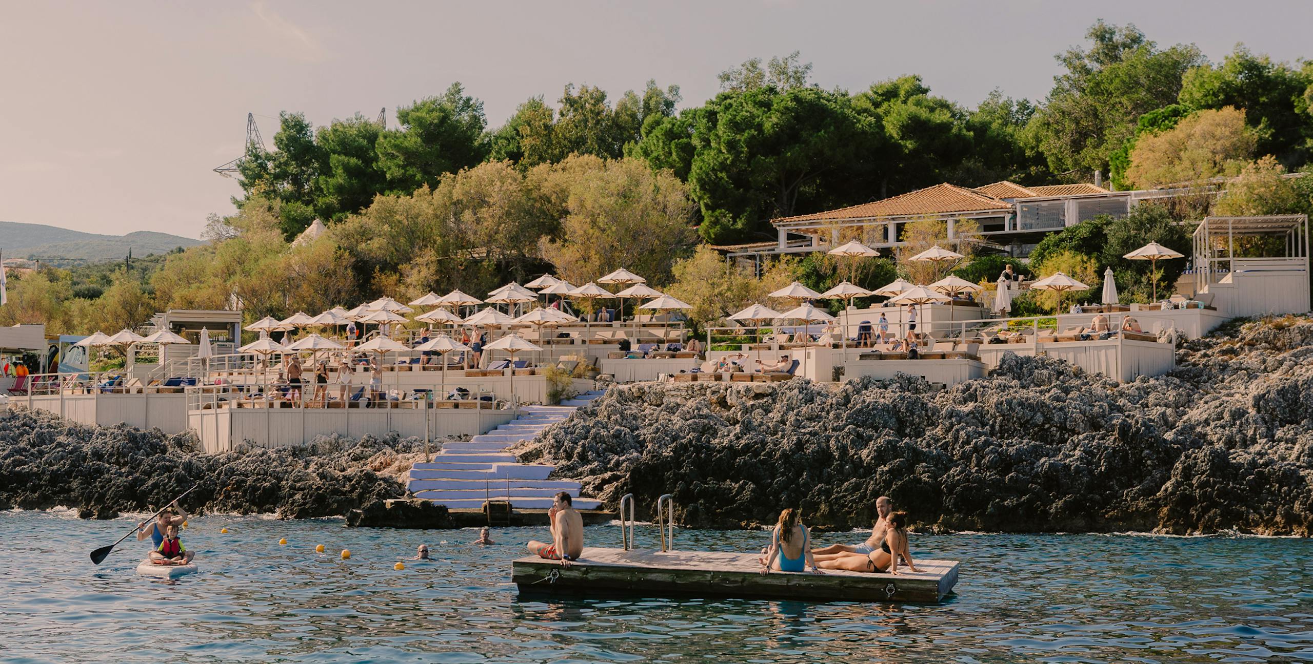 Coastal Mediterranean resort with swimmers and sunbathers enjoying turquoise waters, terraced seating areas with umbrellas, and hillside buildings amid greenery under soft evening light.