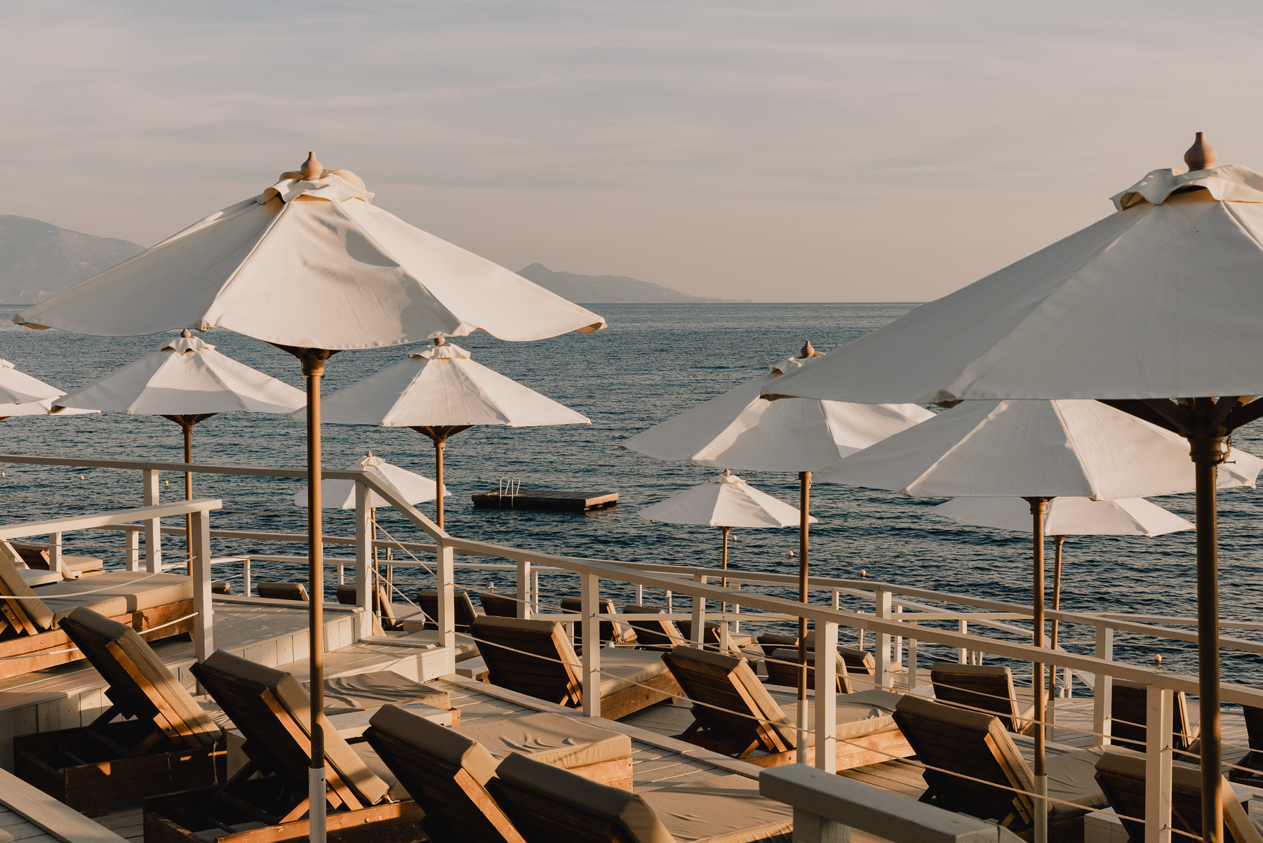 Waterfront sun deck at golden hour featuring rows of wooden loungers with charcoal cushions under billowing white canvas umbrellas with knotted tops, white railings along the water's edge, floating swimming platform in the calm Ionian Sea, and distant misty mountain silhouettes under a soft pastel sky.