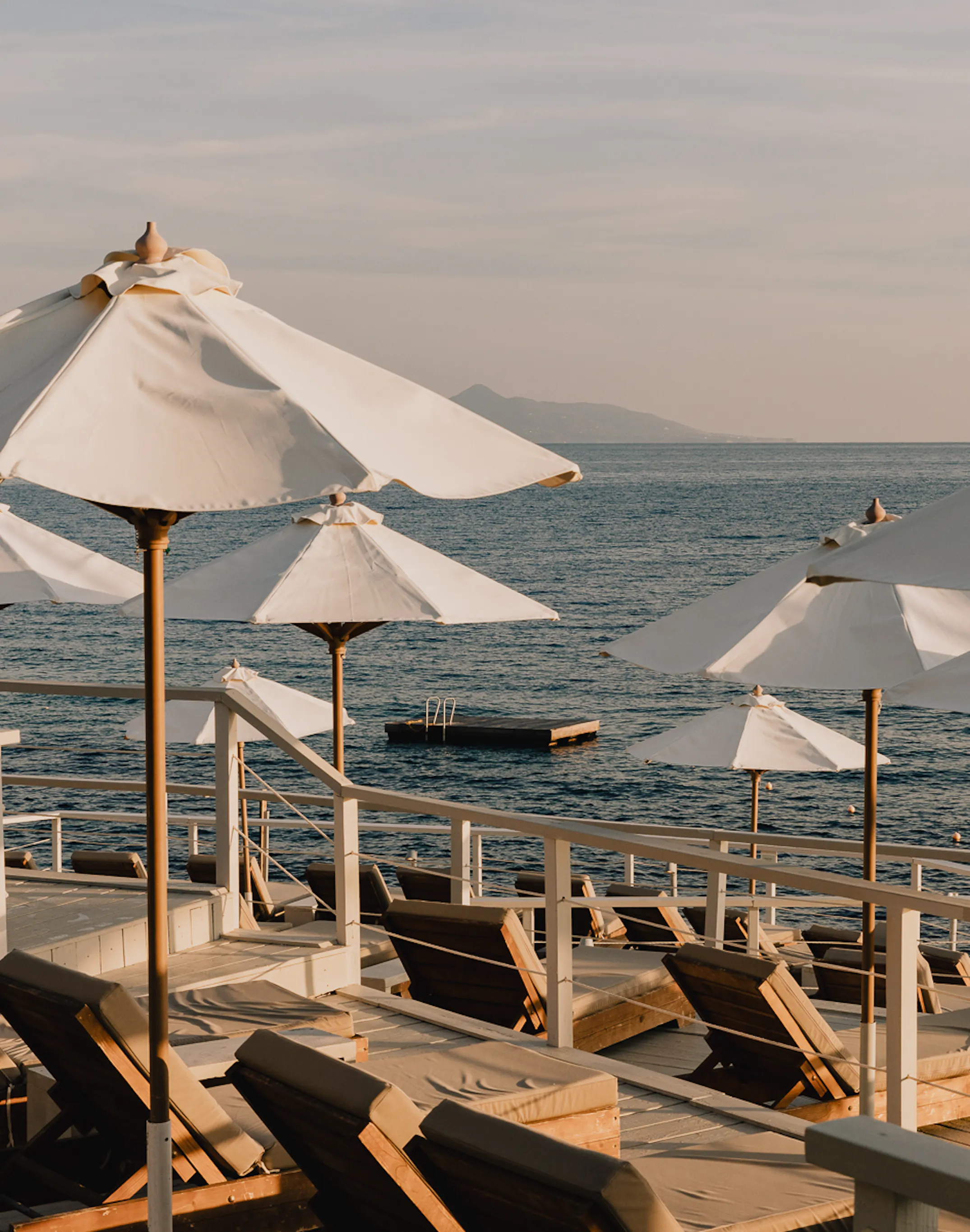 Waterfront sun deck at golden hour featuring rows of wooden loungers with charcoal cushions under billowing white canvas umbrellas with knotted tops, white railings along the water's edge, floating swimming platform in the calm Ionian Sea, and distant misty mountain silhouettes under a soft pastel sky.