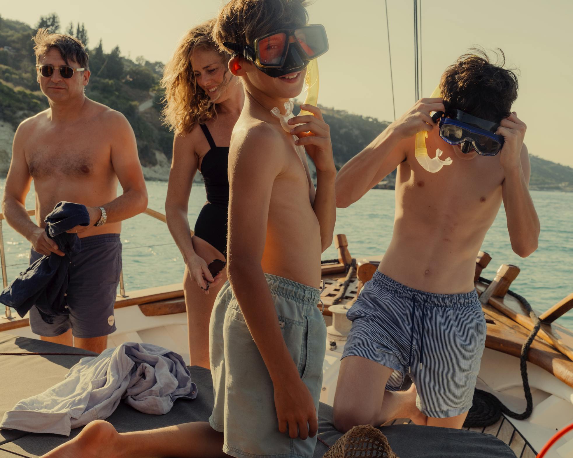 Children prepare to snorkel from a sailboat deck, wearing masks and swim gear, with calm blue waters and green hillsides beyond—embodying carefree summer adventure and watersports activities.
