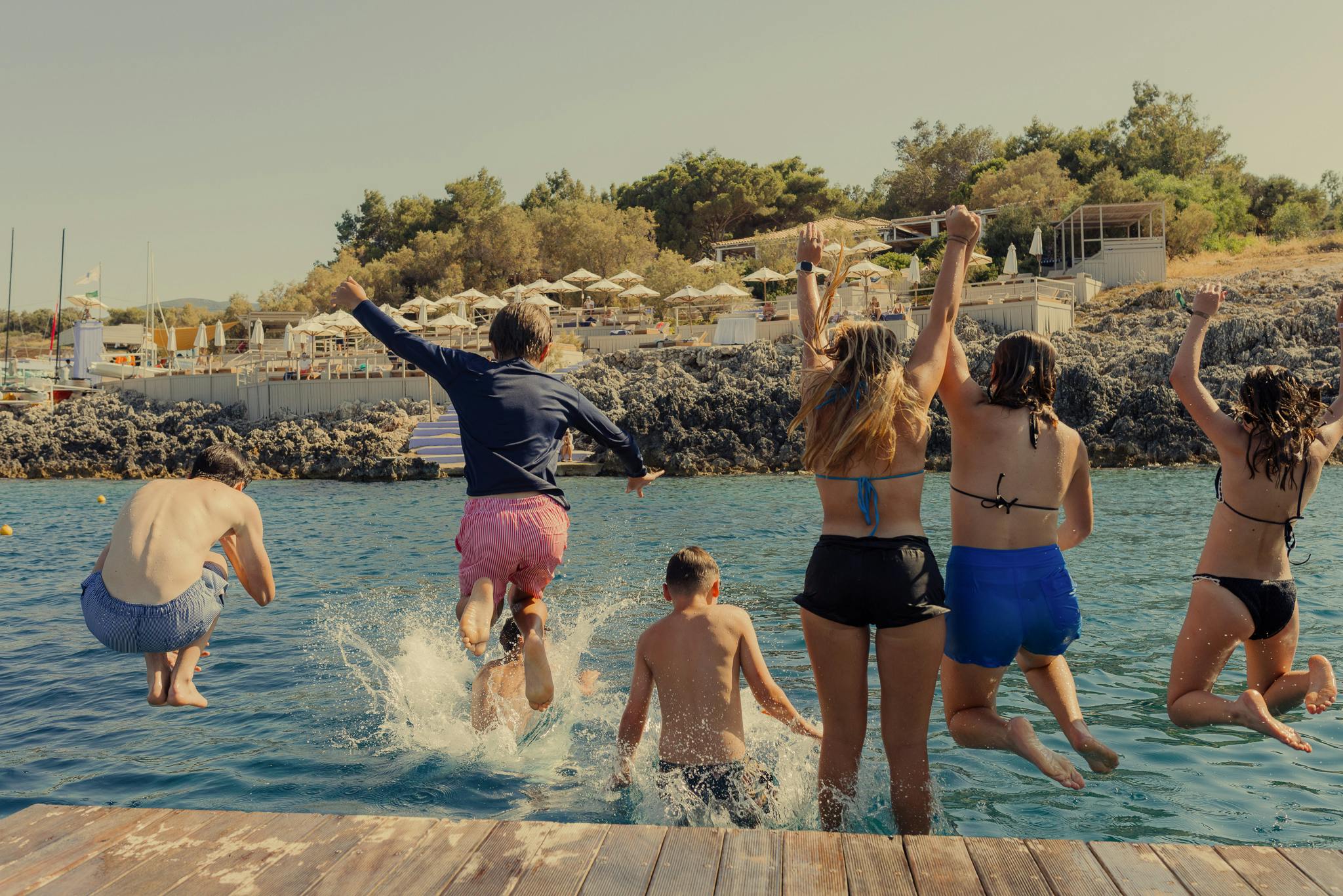 A group of teenagers and children leap simultaneously from a wooden dock into turquoise waters, with the beach club's rocky shore and sun loungers visible beyond—capturing pure joy and spontaneous summer fun.