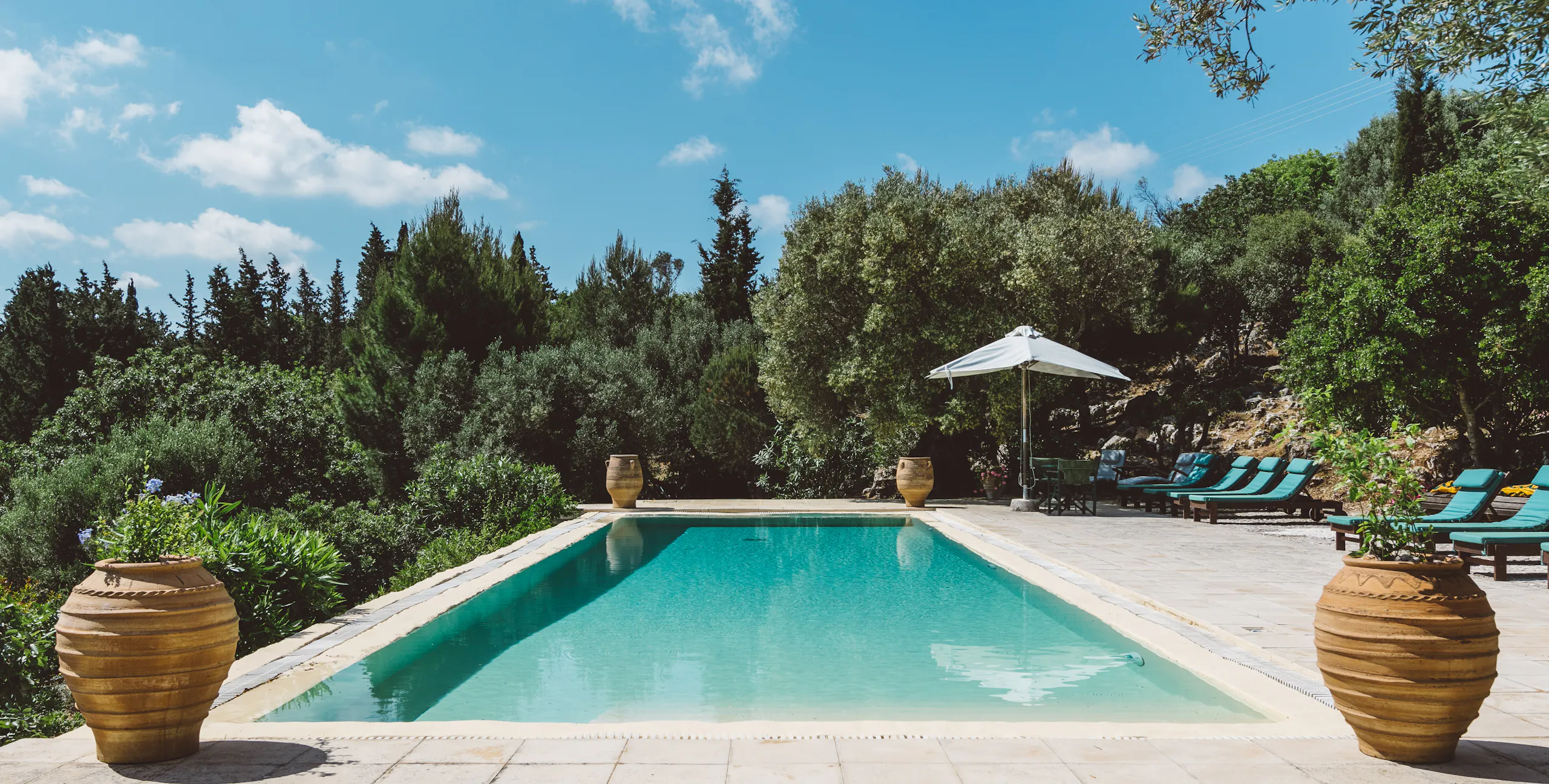 A rectangular outdoor swimming pool with clear turquoise water, surrounded by a stone patio. Two large terracotta urns flank the foreground. Sun loungers and a white umbrella are positioned poolside, with lush green trees and shrubs in the background under a bright blue sky.