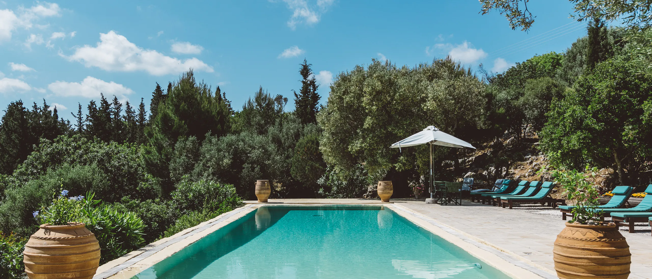 A rectangular outdoor swimming pool with clear turquoise water, surrounded by a stone patio. Two large terracotta urns flank the foreground. Sun loungers and a white umbrella are positioned poolside, with lush green trees and shrubs in the background under a bright blue sky.
