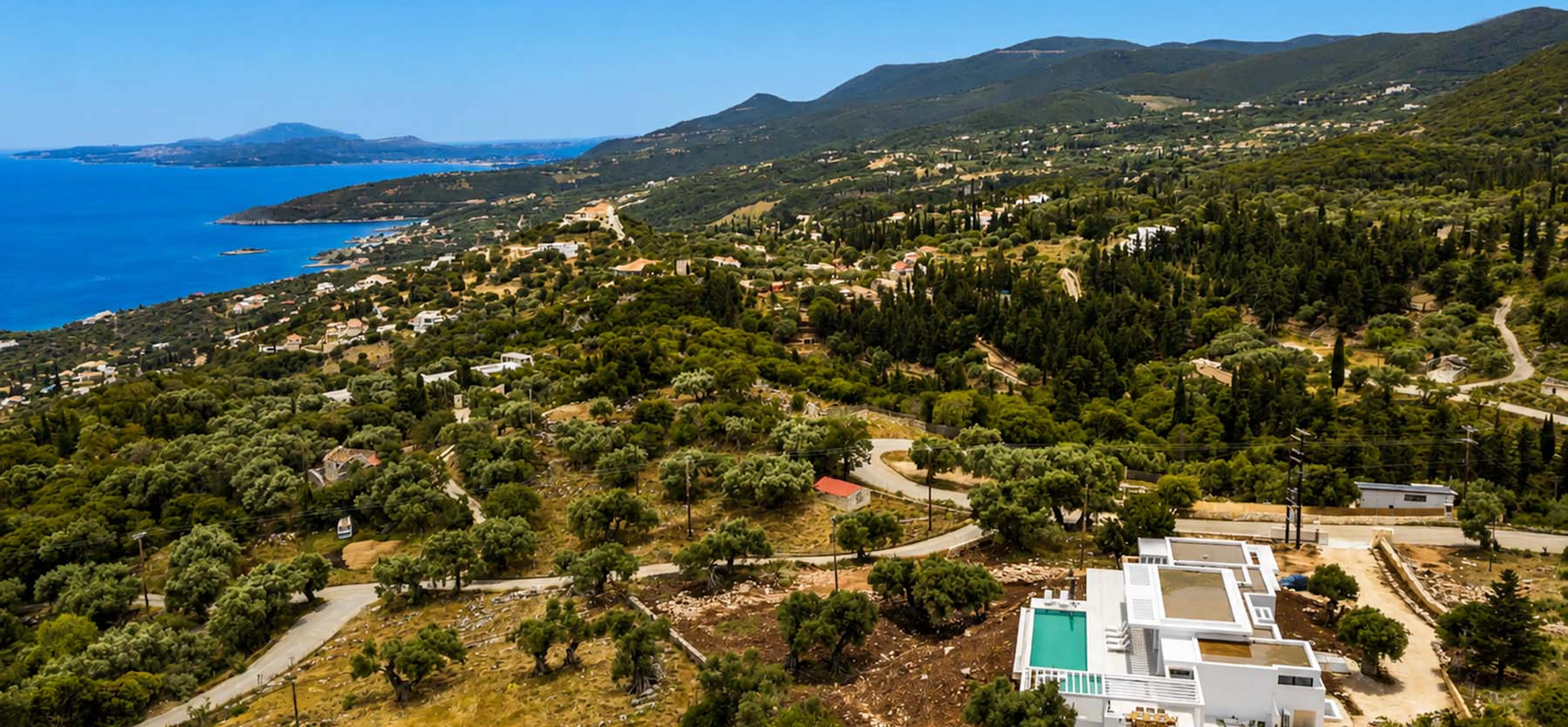 Aerial drone view of a white contemporary villa with swimming pool set among olive groves on a hillside, with sweeping views across the coastline and deep blue sea