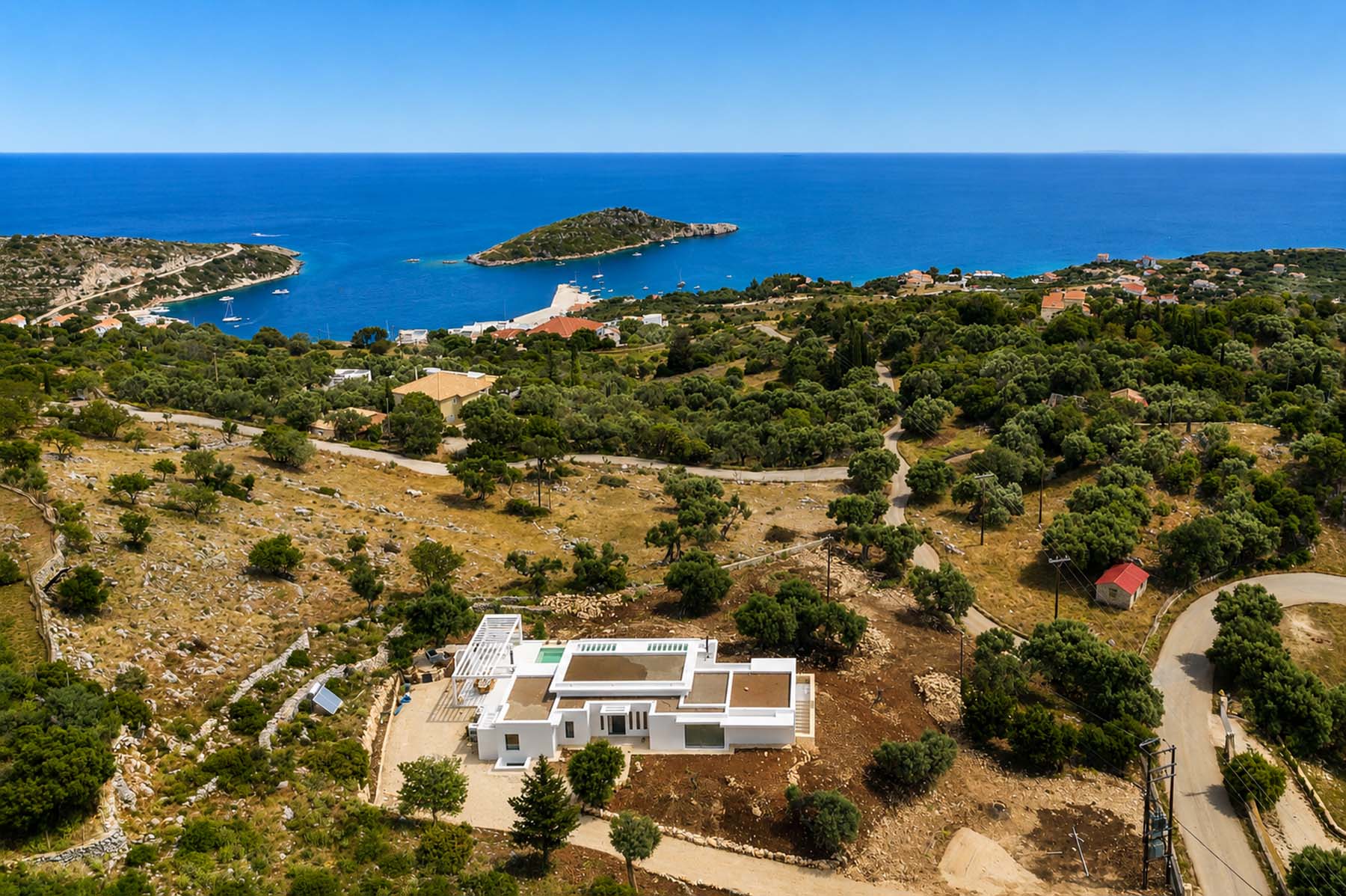 Aerial view showing the white villa and its pool amid dry olive groves, with a sheltered bay, small island, and moored sailing boats visible in the vivid blue sea behind
