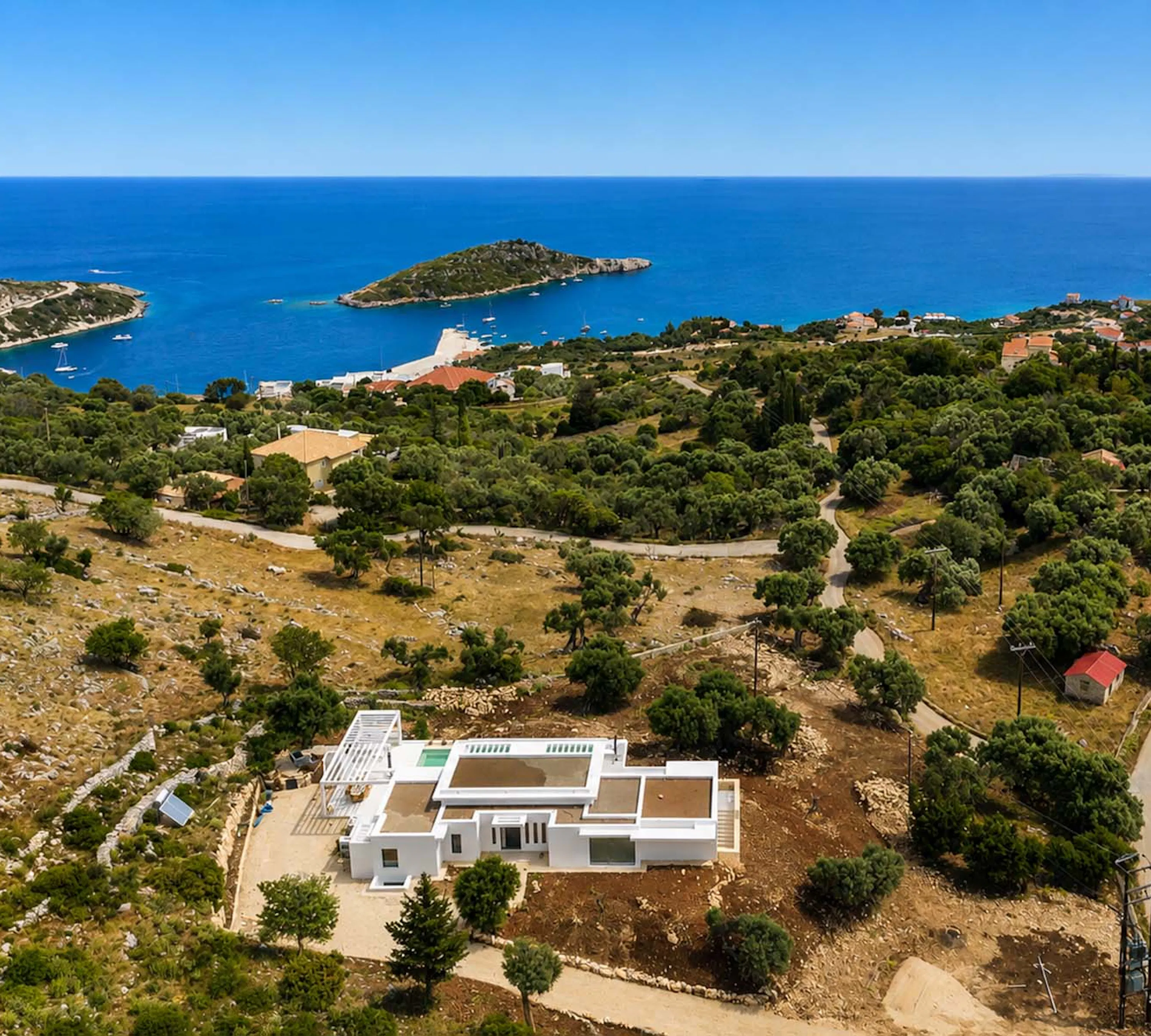 Aerial view showing the white villa and its pool amid dry olive groves, with a sheltered bay, small island, and moored sailing boats visible in the vivid blue sea behind