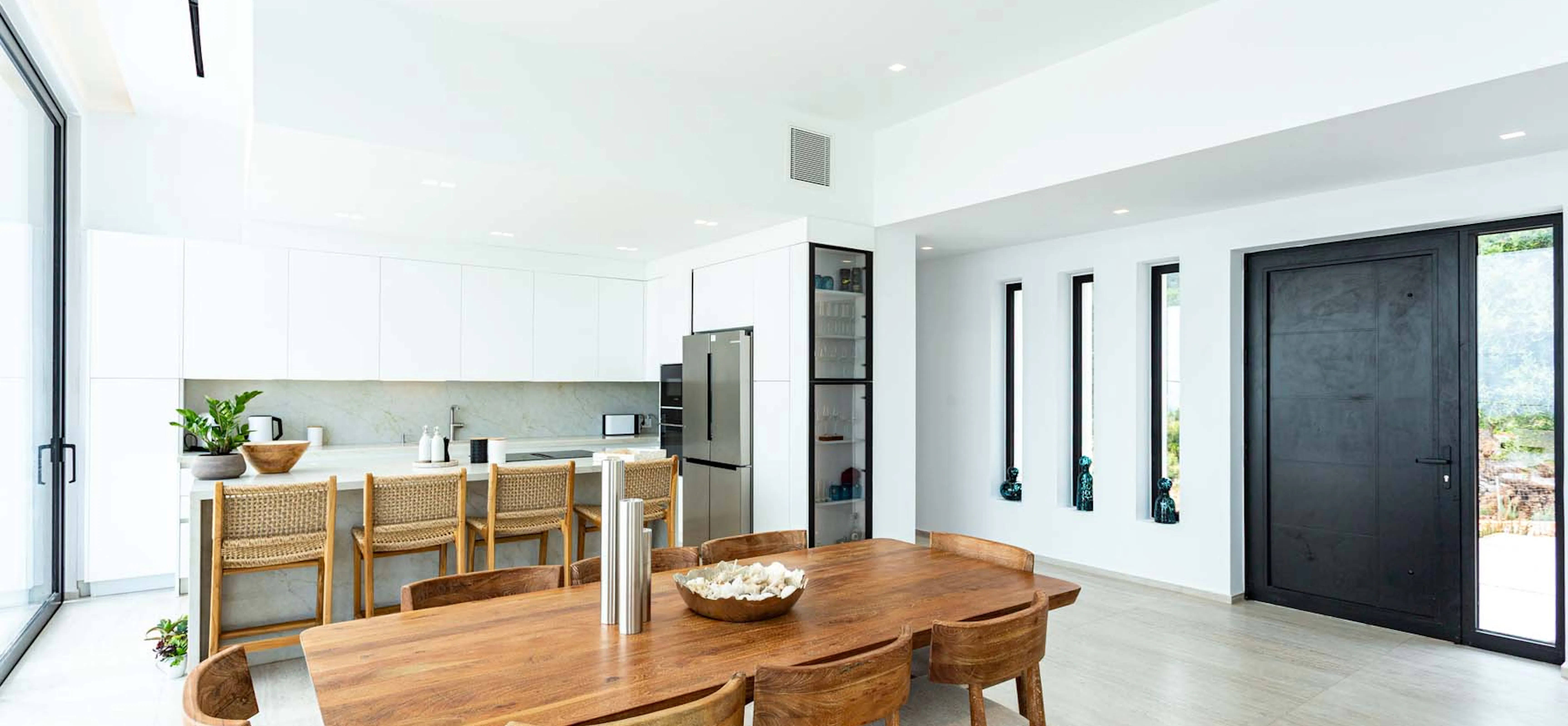 Bright open-plan kitchen and dining area with a solid wood dining table and sculpted wooden chairs, rattan bar stools at a stone island, white handleless cabinetry, stainless steel fridge, and a tall black front door flanked by narrow vertical windows