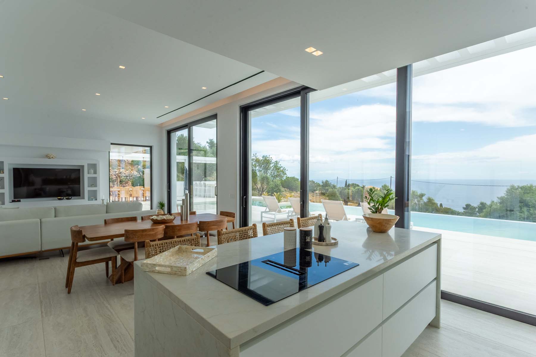 Open-plan kitchen and living space with a white induction island, wooden dining table and rattan chairs, a large sectional sofa, built-in TV wall unit, and floor-to-ceiling black-framed glass doors opening directly onto the pool terrace and sea view beyond
