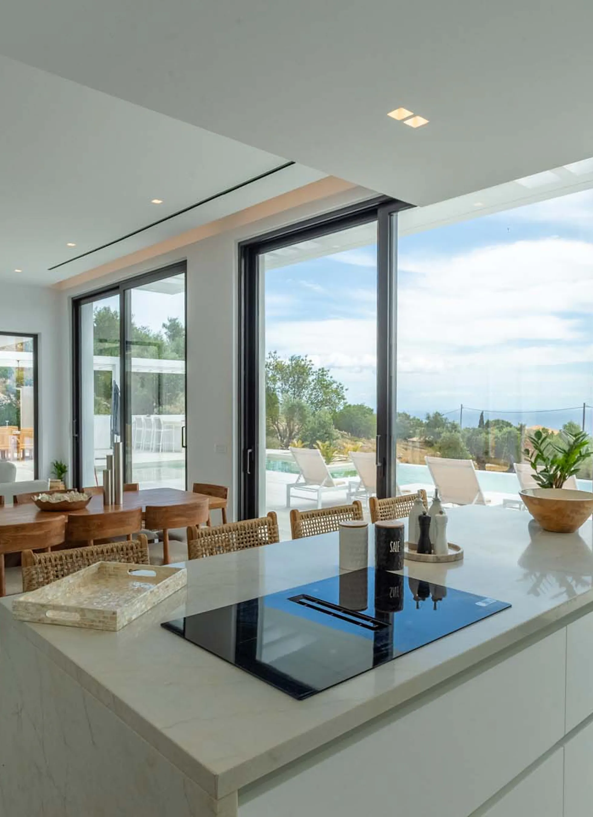 Open-plan kitchen and living space with a white induction island, wooden dining table and rattan chairs, a large sectional sofa, built-in TV wall unit, and floor-to-ceiling black-framed glass doors opening directly onto the pool terrace and sea view beyond