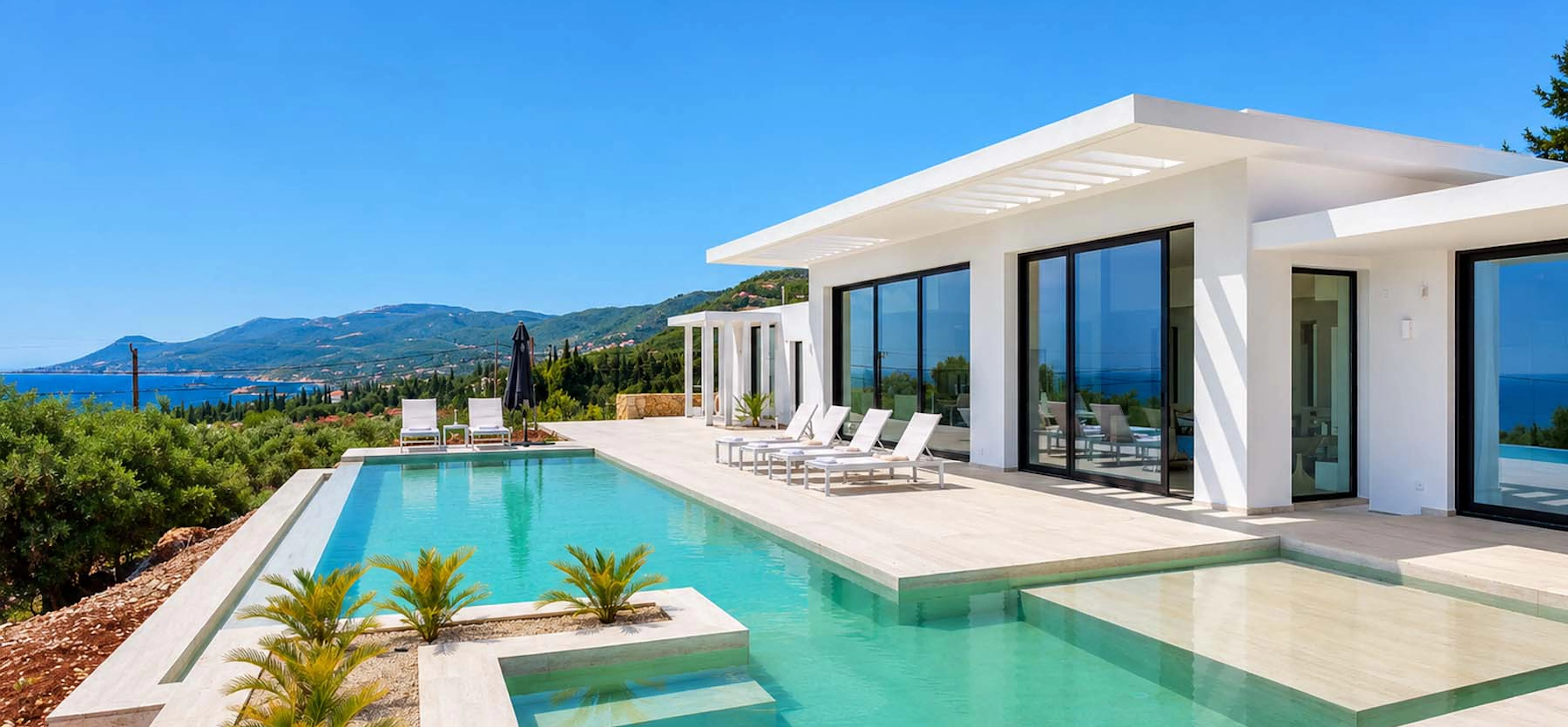 White modernist villa with floor-to-ceiling black-framed glass doors, a stepped infinity pool with flanking wading area and tropical plantings, white sun loungers on a stone terrace, and panoramic views across the olive-covered hillside to the sea and mountains
