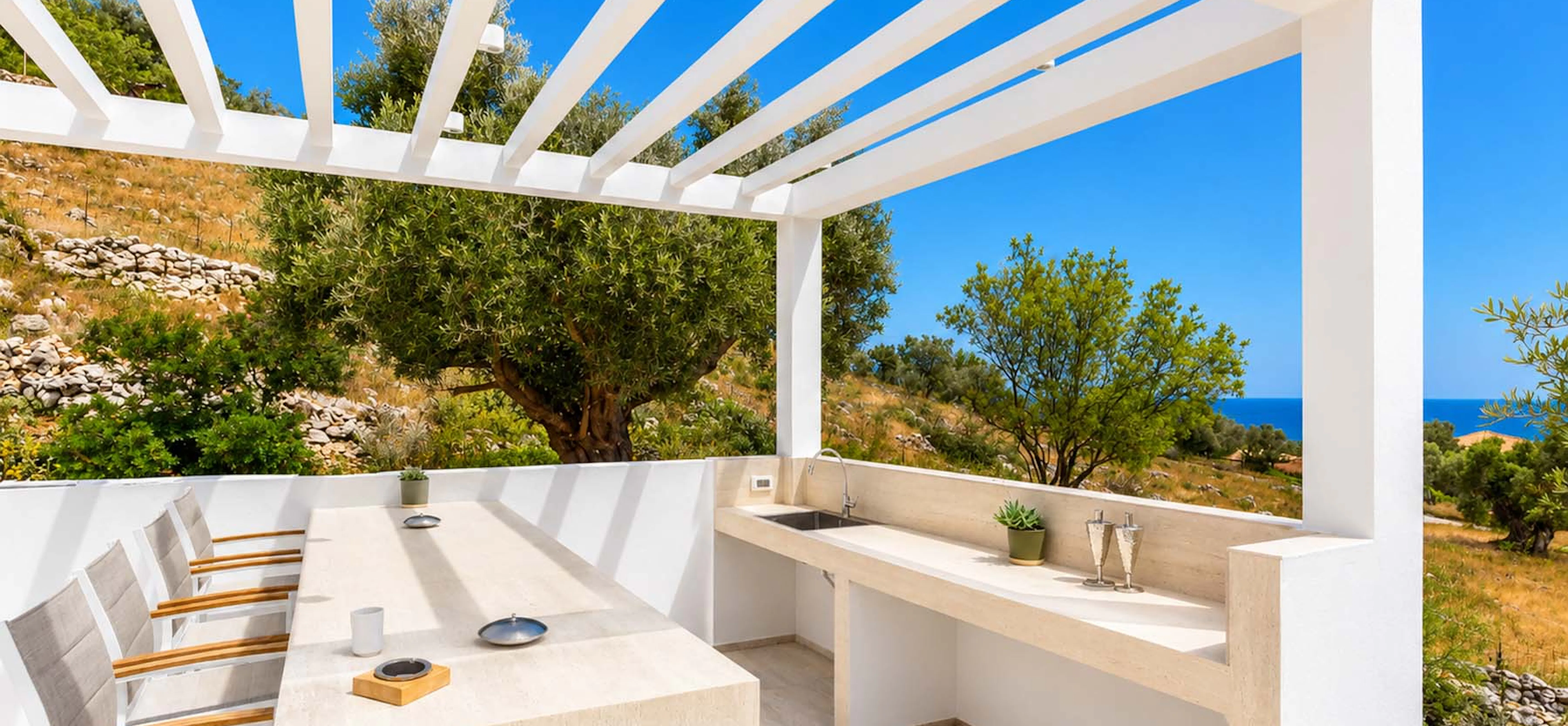 Covered outdoor dining terrace with a white pergola, stone-topped built-in kitchen counter, teak and aluminium dining chairs, framed by olive trees and a sea view under a deep blue sky