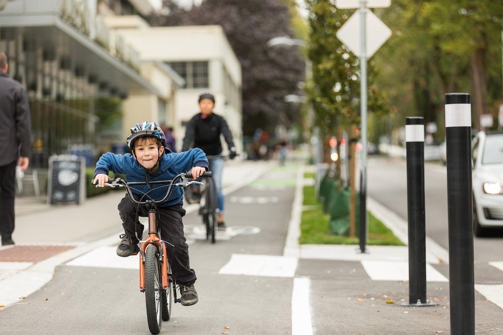 A young, elementary school aged child rides an orange bike in a protected bike lane. Their parent rides behind them. 