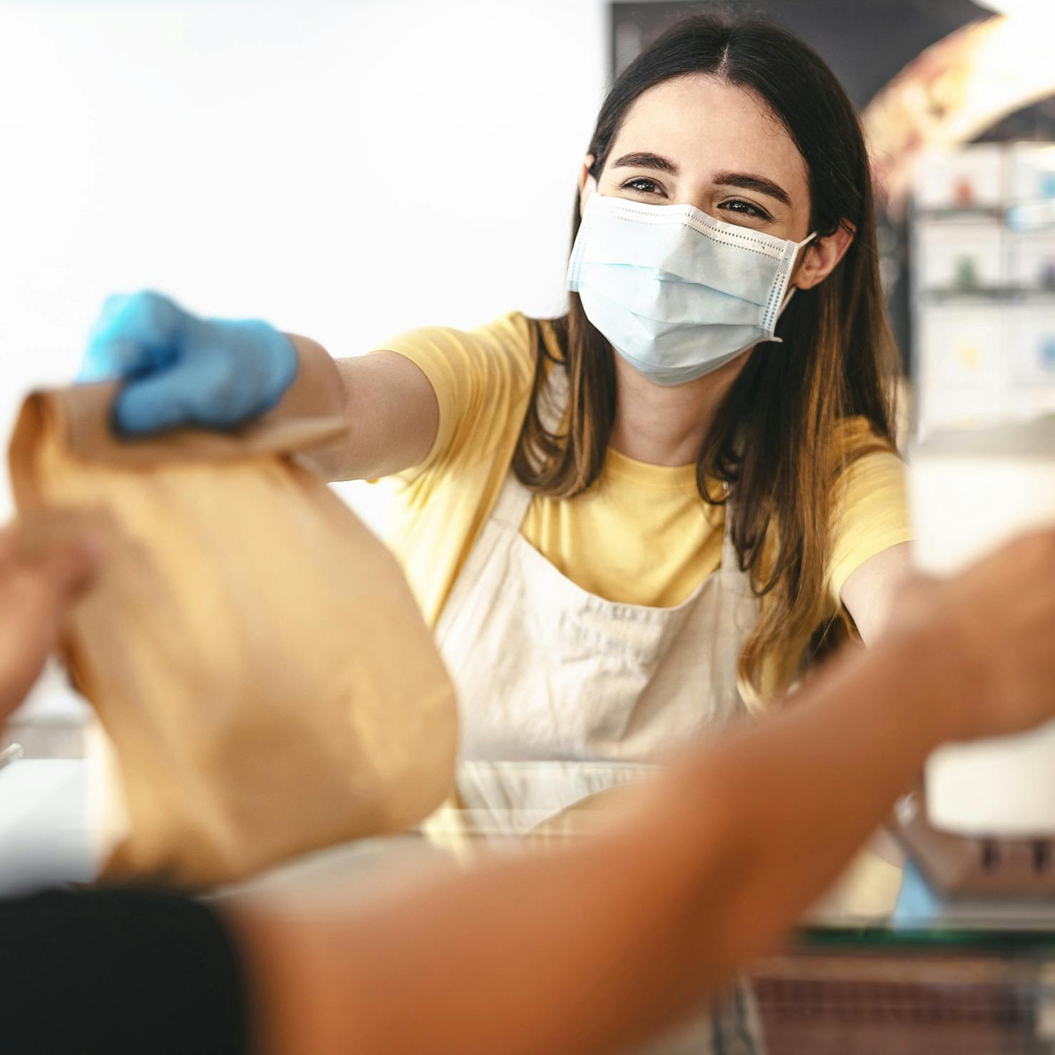 Woman wearing mask and gloves, handing a bag of food to customer.