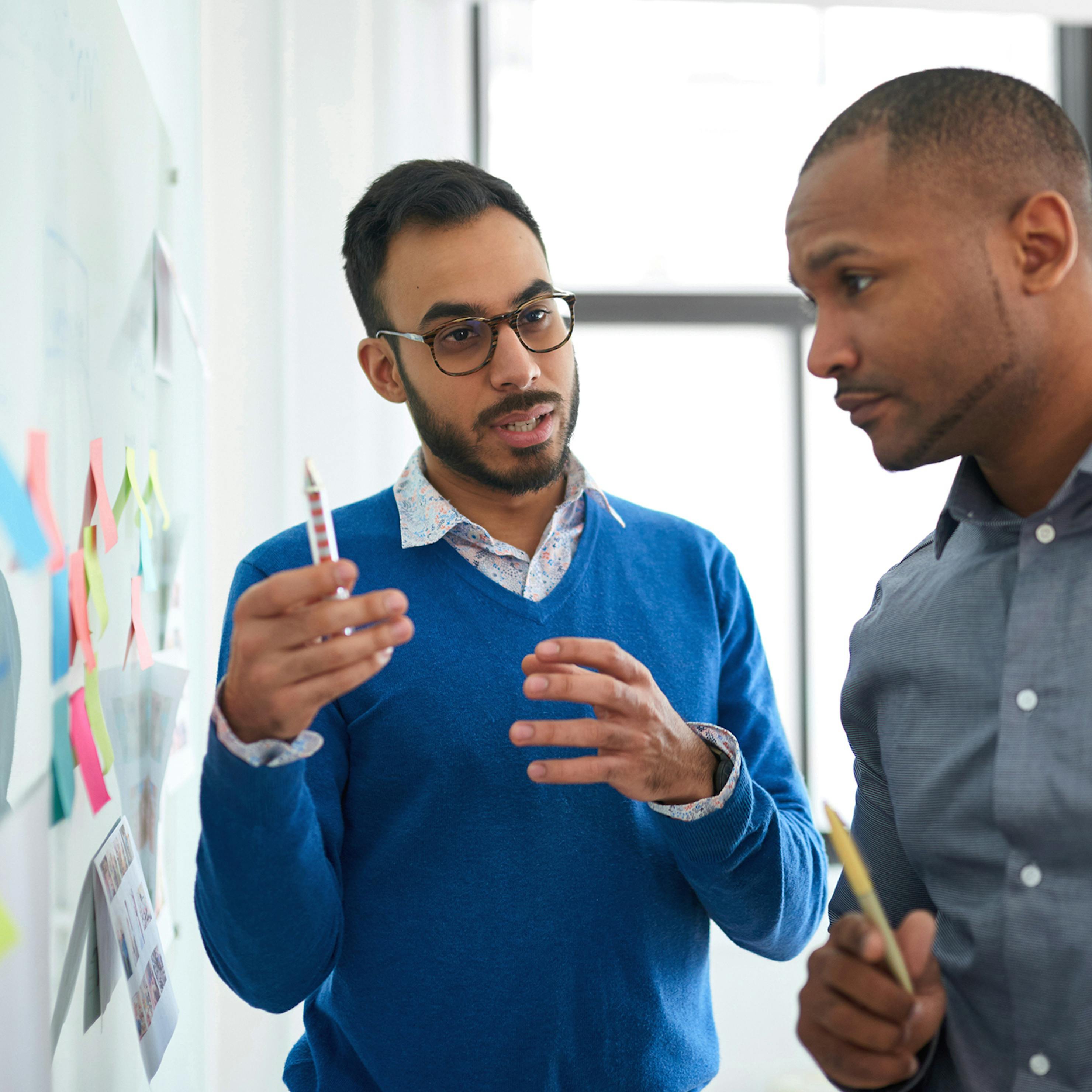 Man wearing glasses and holding pen while speaking to a colleague in front of a white board with stickies.