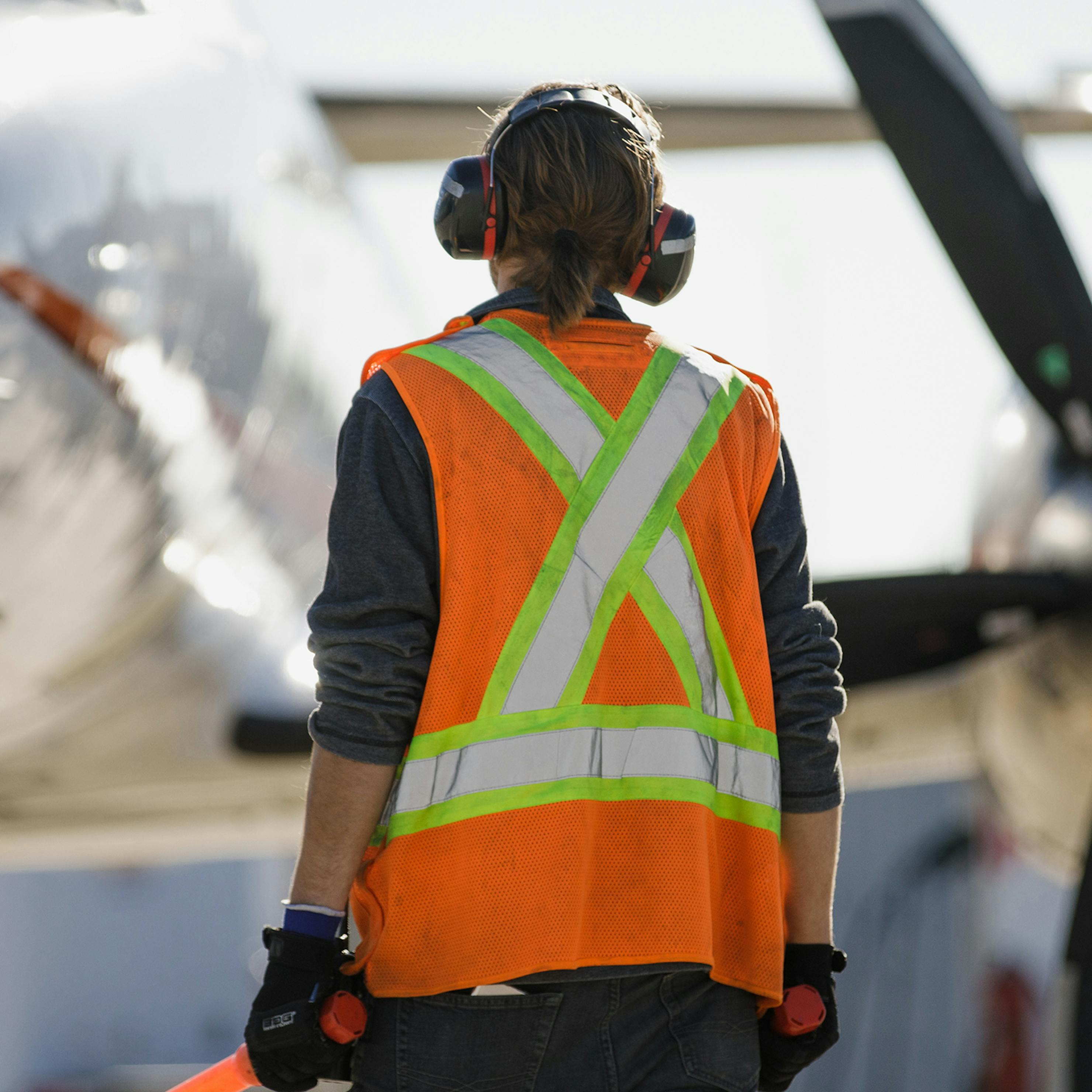 Man standing on apron holding orange batons and wearing hearing protection. Ready to direct the aircraft.