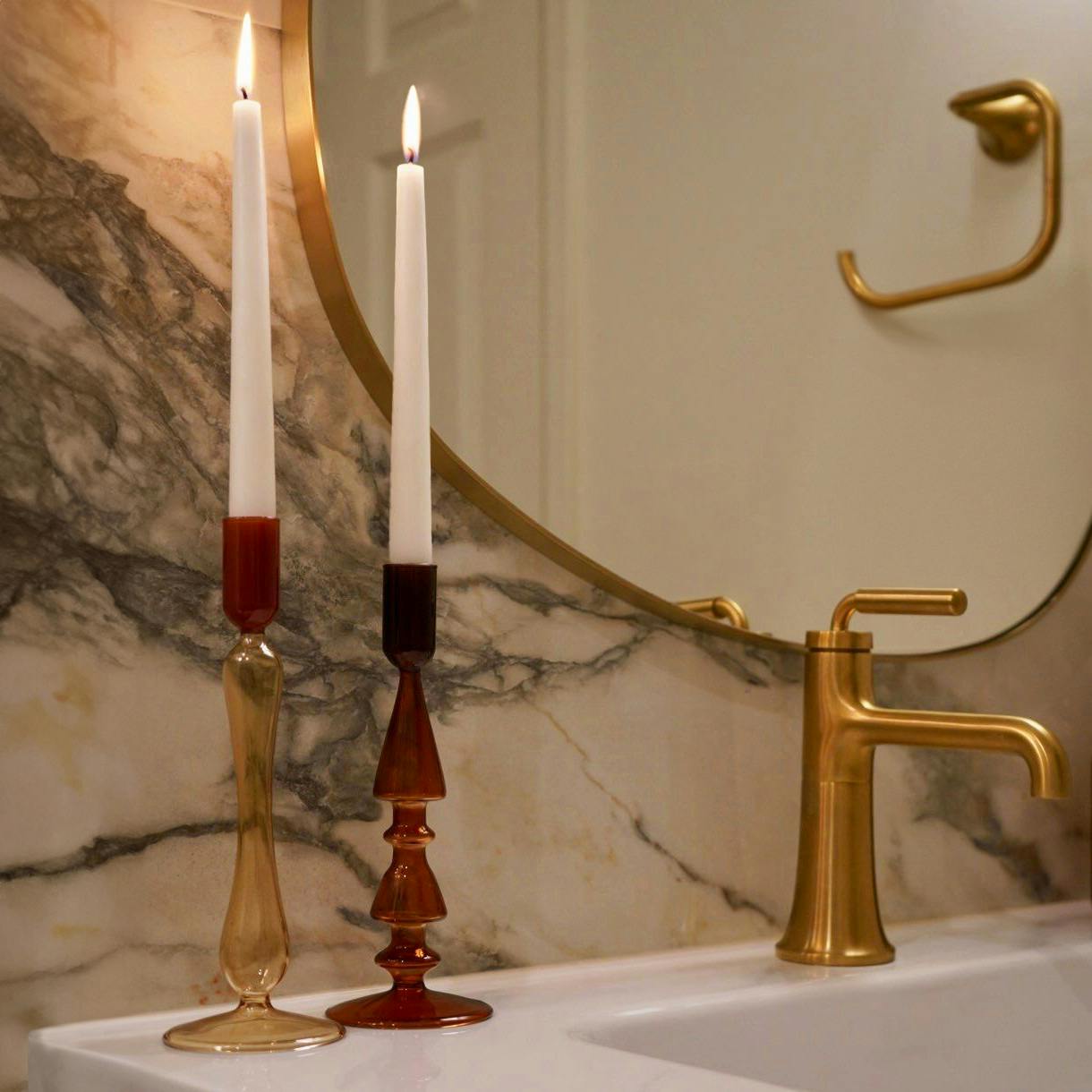 This bathroom exudes quiet luxury with richly veined marble and warm brass accents. A pair of sculptural candlesticks and a softly curved mirror elevate the space with a sense of timeless elegance.