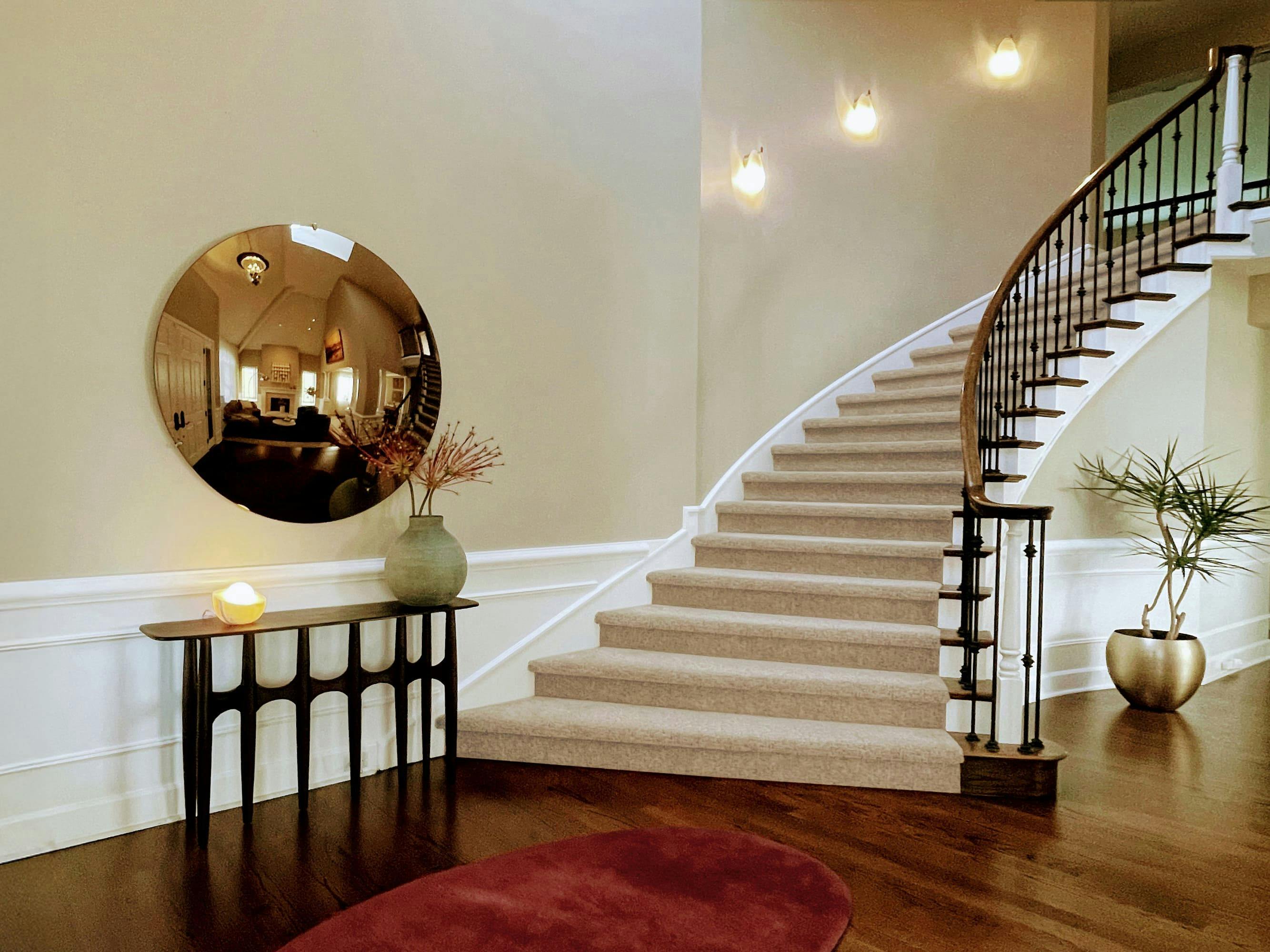 In this Entry Foyer designed by Persimmon Design, a sculptural smoked bronze mirror greets with a warm glimpse into the seating area beyond. Paired with the sweep of the staircase and a slender console, it sets a refined and inviting tone from the moment you step inside the home.