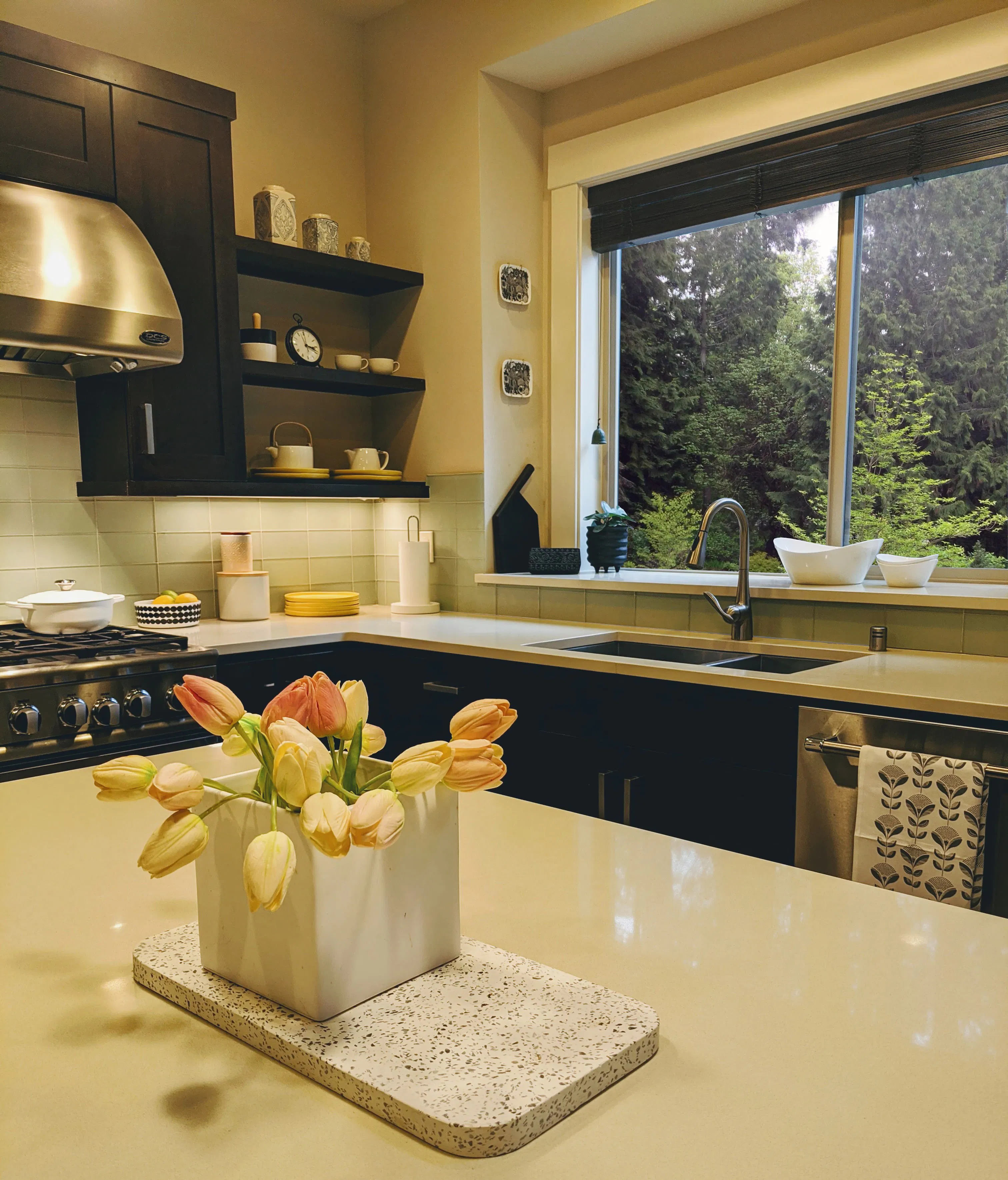 In this Pacific Northwest kitchen by Persimmon Design, the calm, cohesive atmosphere comes from a carefully developed design concept that balances warmth, functionality, and a strong connection to the surrounding landscape. The expansive window framing the trees, layered lighting, open shelving displaying the client’s treasured objects, and restrained palette work together to create a kitchen that feels grounded, welcoming, and unmistakably personal.