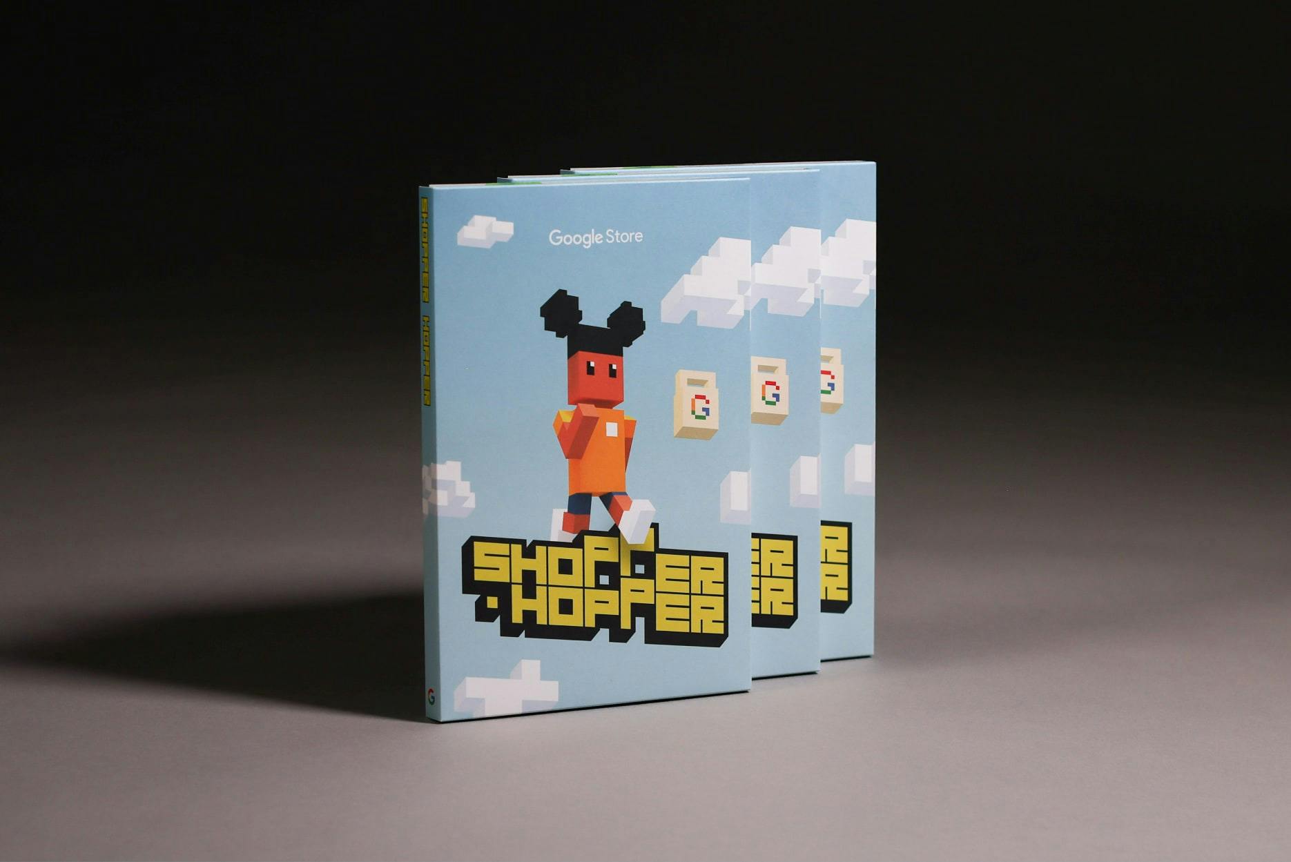 Studio shot of three product boxes standing upright, slightly fanned out. The boxes feature artwork for the Google Store game 'Shopper Hopper,' showing a blocky, voxel-art character walking above the game's yellow and black logo. The background is a light blue sky with pixelated white clouds.