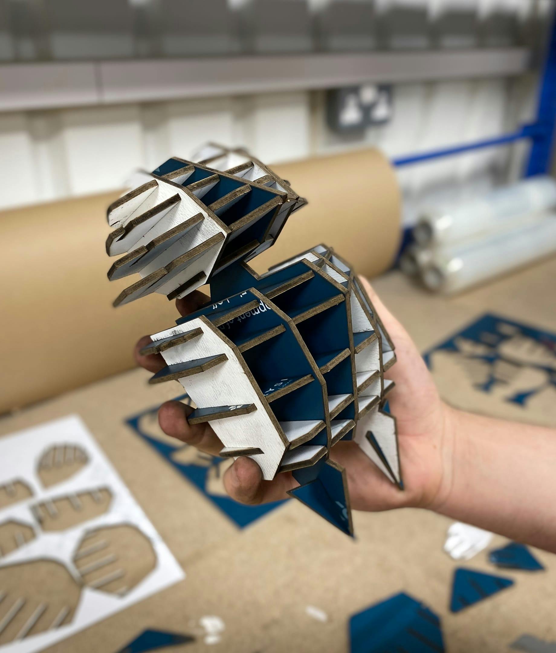 A person's hand holds up a 3D geometric character, partially assembled from slotted pieces of cardboard. The outer surfaces are white, while the inner surfaces are blue. In the background, on a workshop table, are the leftover cardboard sheets from which the pieces were punched.
