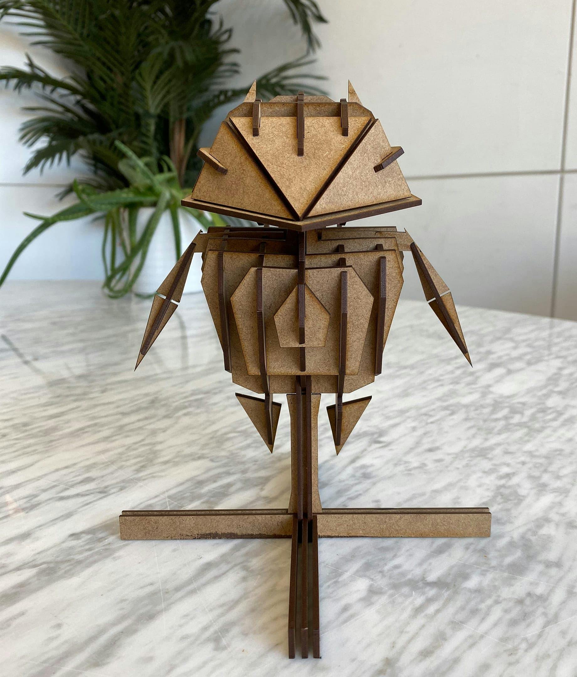 A 3D model of a geometric character, assembled from interlocking pieces of laser-cut wood or MDF. The character resembles a robot with a large head and pointed ears, and it stands on a cross-shaped base. The model is unpainted, showing the natural brown color of the material. It is placed on a white and grey marble countertop, with a leafy green plant in a pot visible in the blurred background.