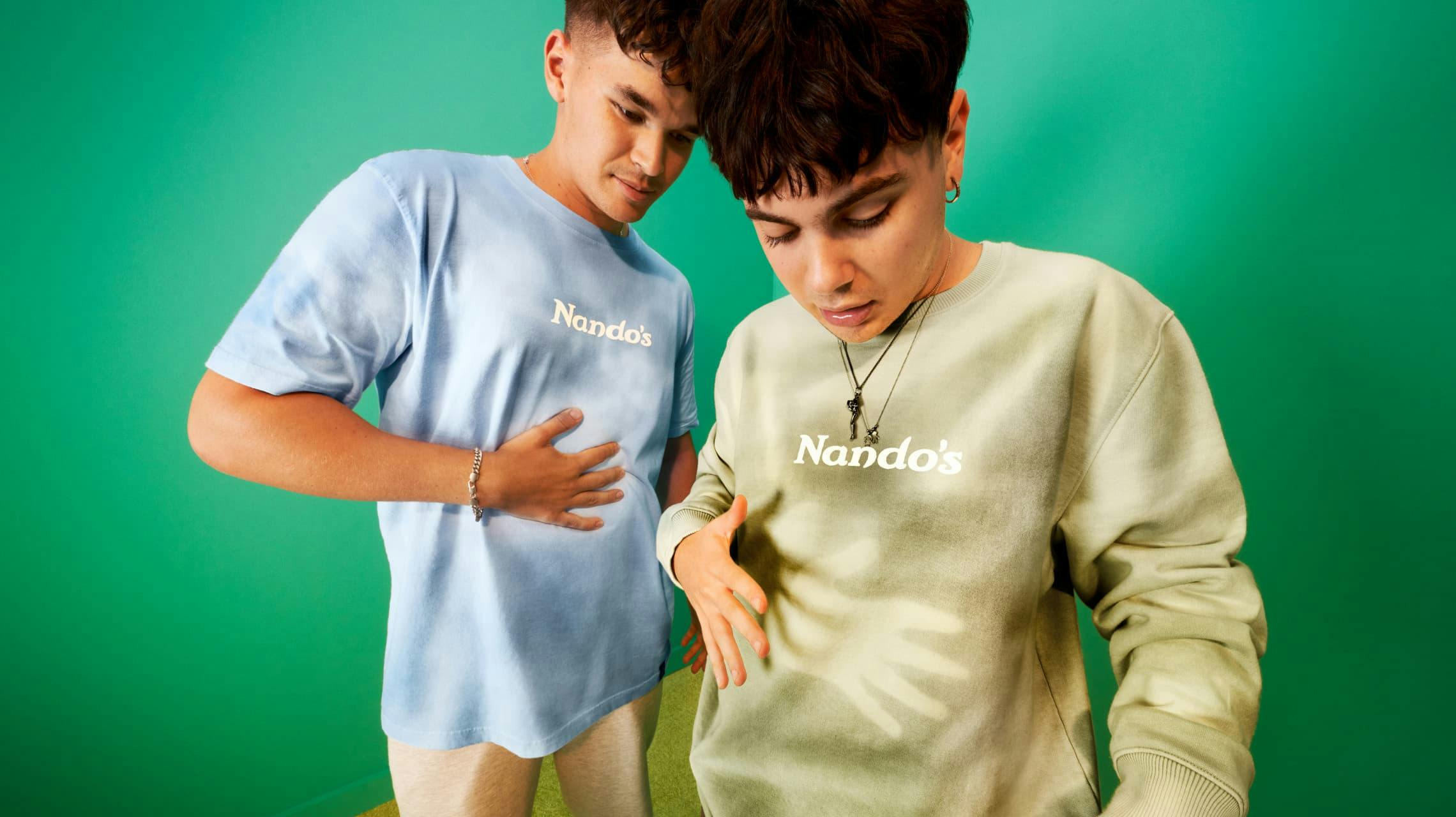Two models posing against a bright green backdrop. One wears a light blue tie-dye Nando's t-shirt and looks down at their stomach. The other, in the foreground, wears a light green Nando's sweatshirt and also looks down, with a shadow of a hand cast on their shirt.